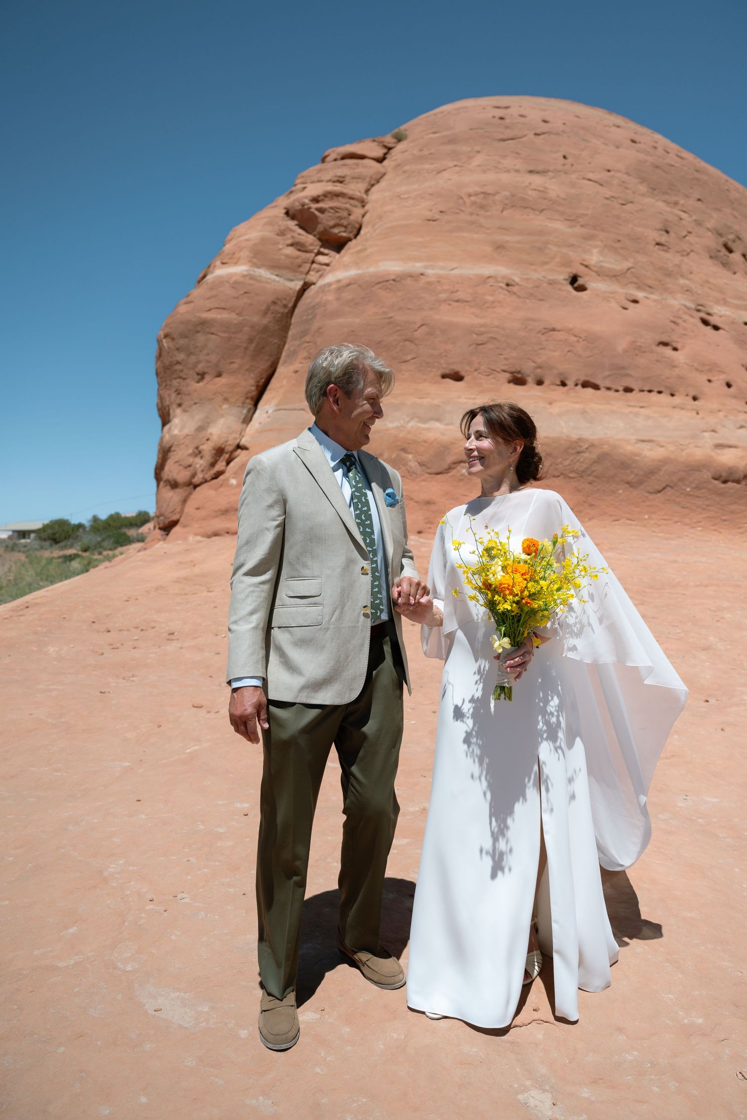 Wedding couple walks hand-in-hand before a massive red sandstone rock formation in the desert.