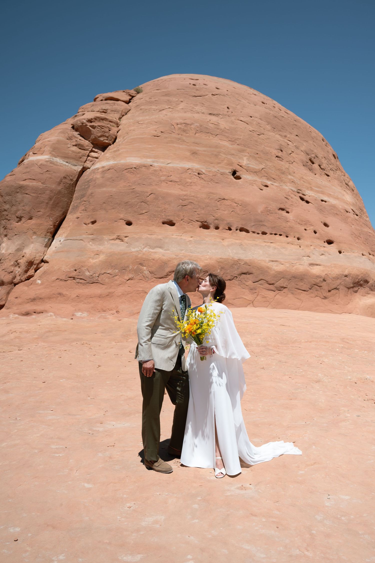 Wedding couple kissing before red rock formation in desert landscape.