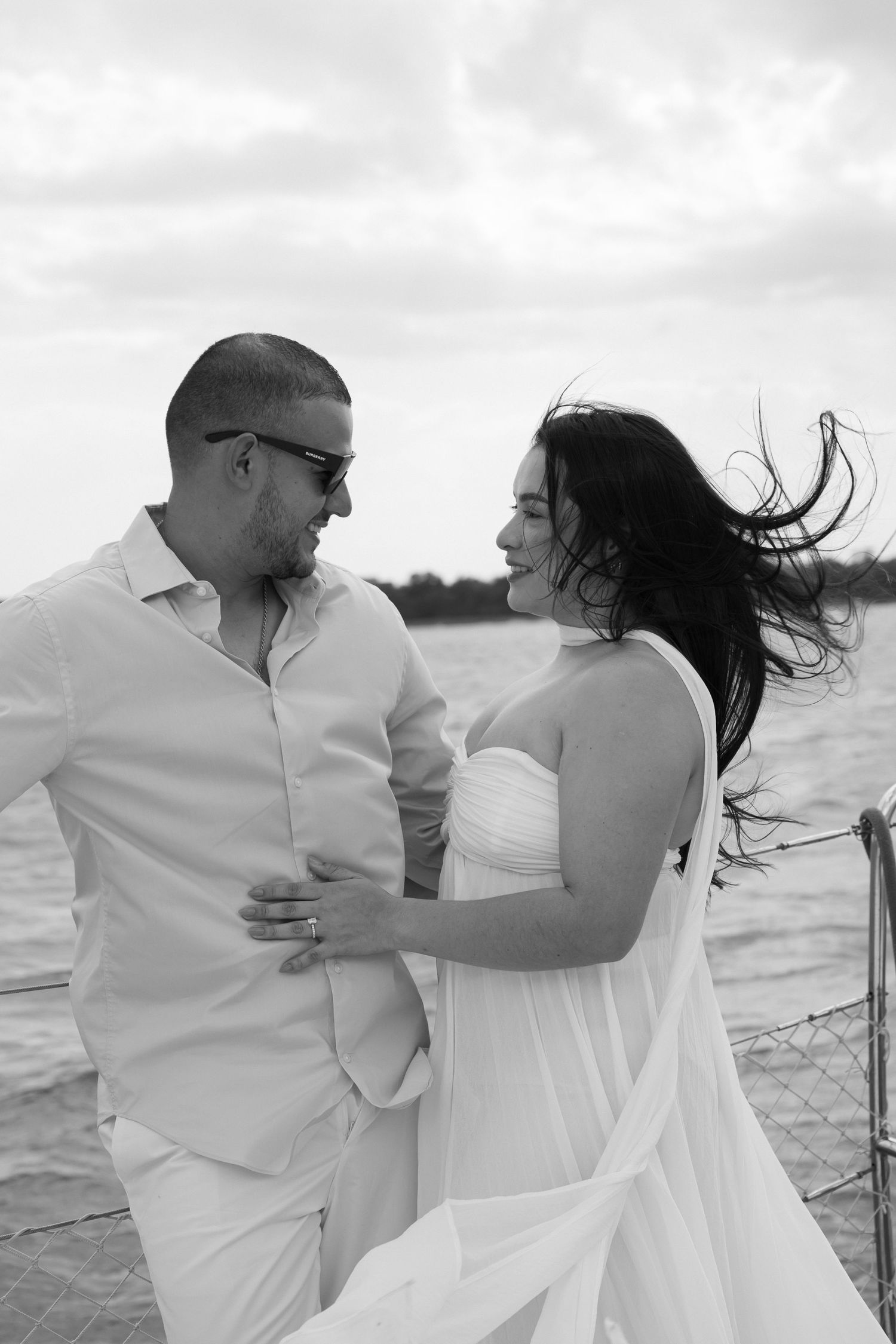 Black and white photo of a couple embracing on a sailboat, woman in white dress with hair blowing in the wind.