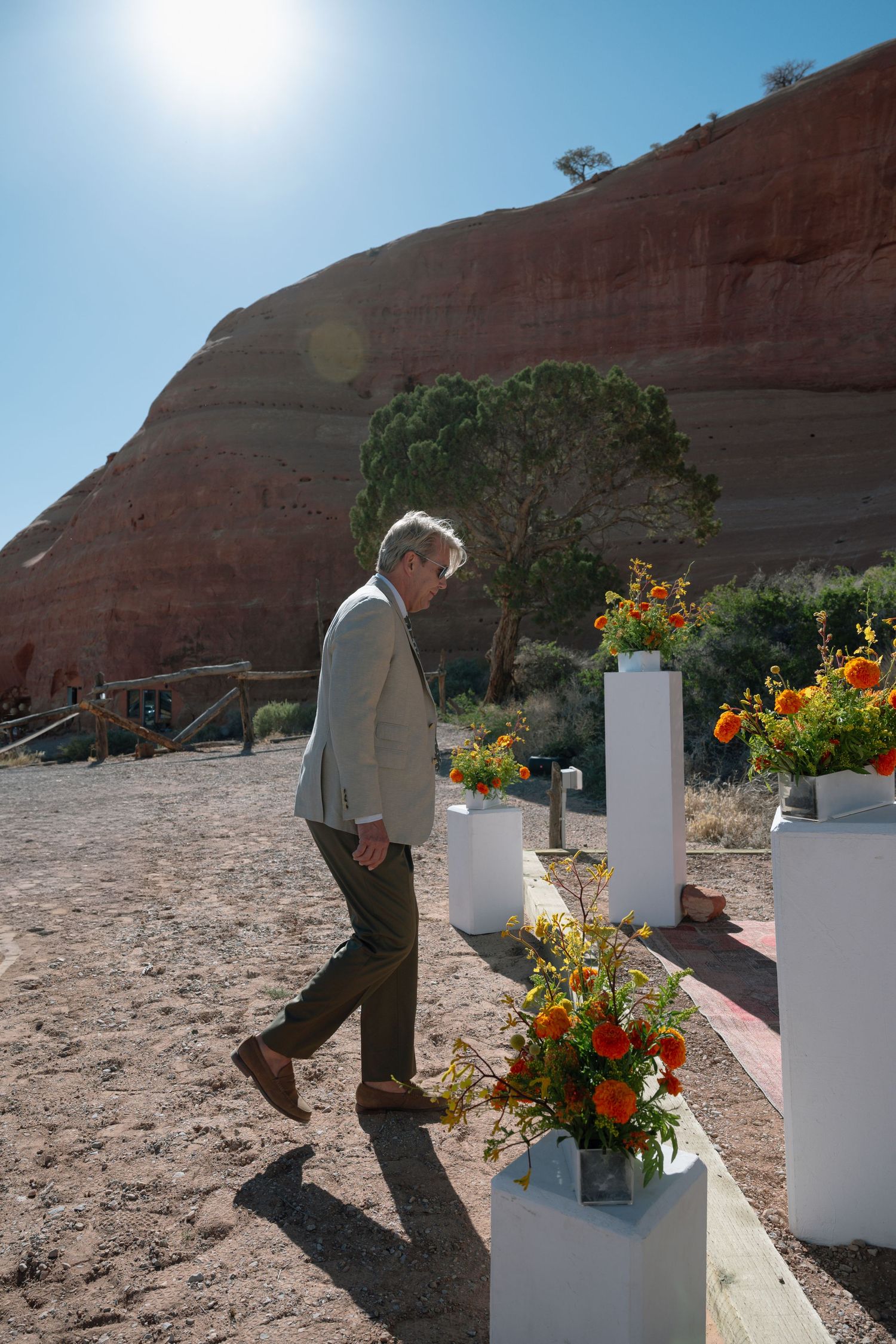 Groom in tan suit walking past white pedestals with colorful floral arrangements at desert wedding venue.