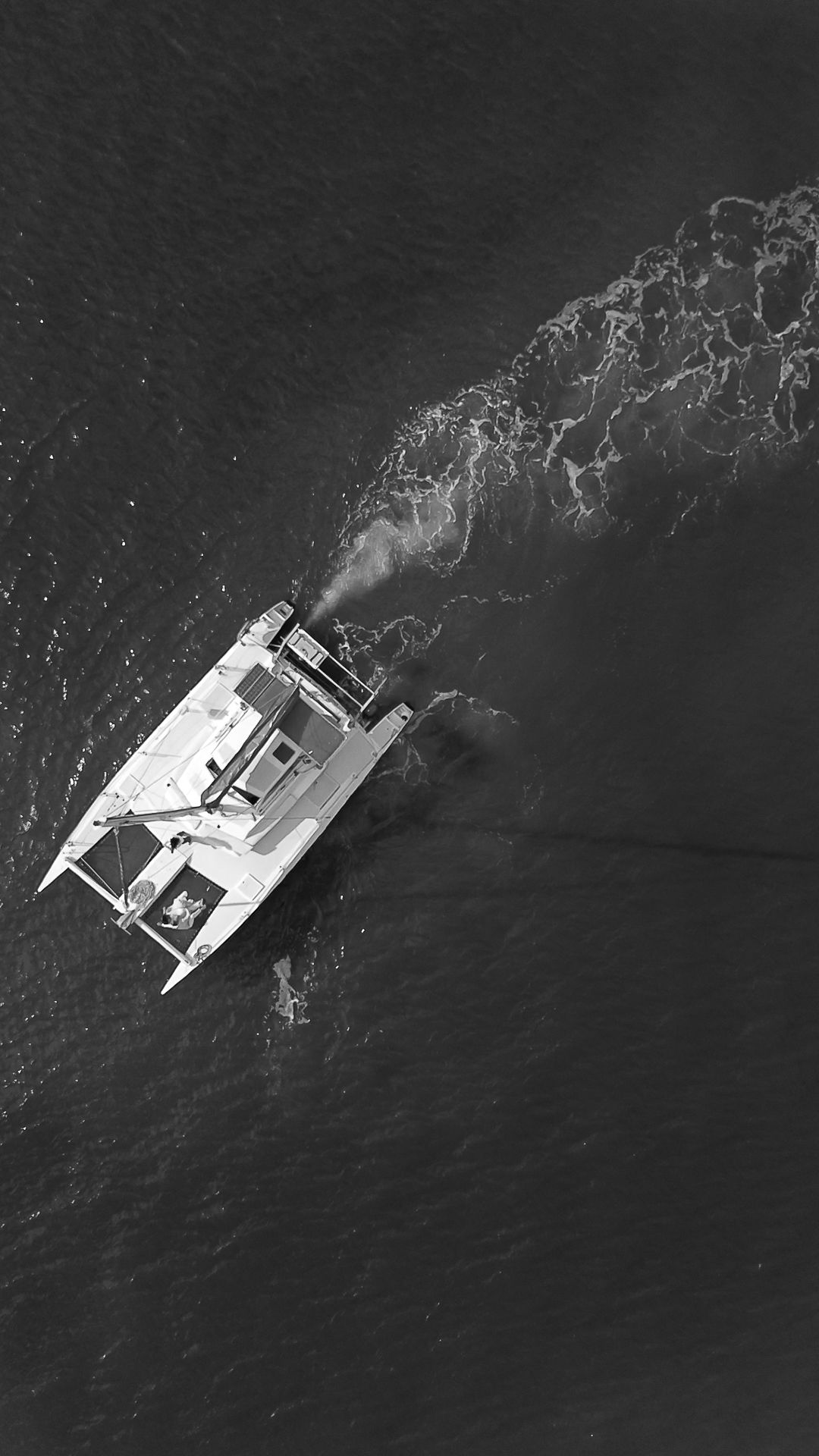 Aerial black and white photo of a catamaran sailing through open ocean waters, leaving a white wake trail.