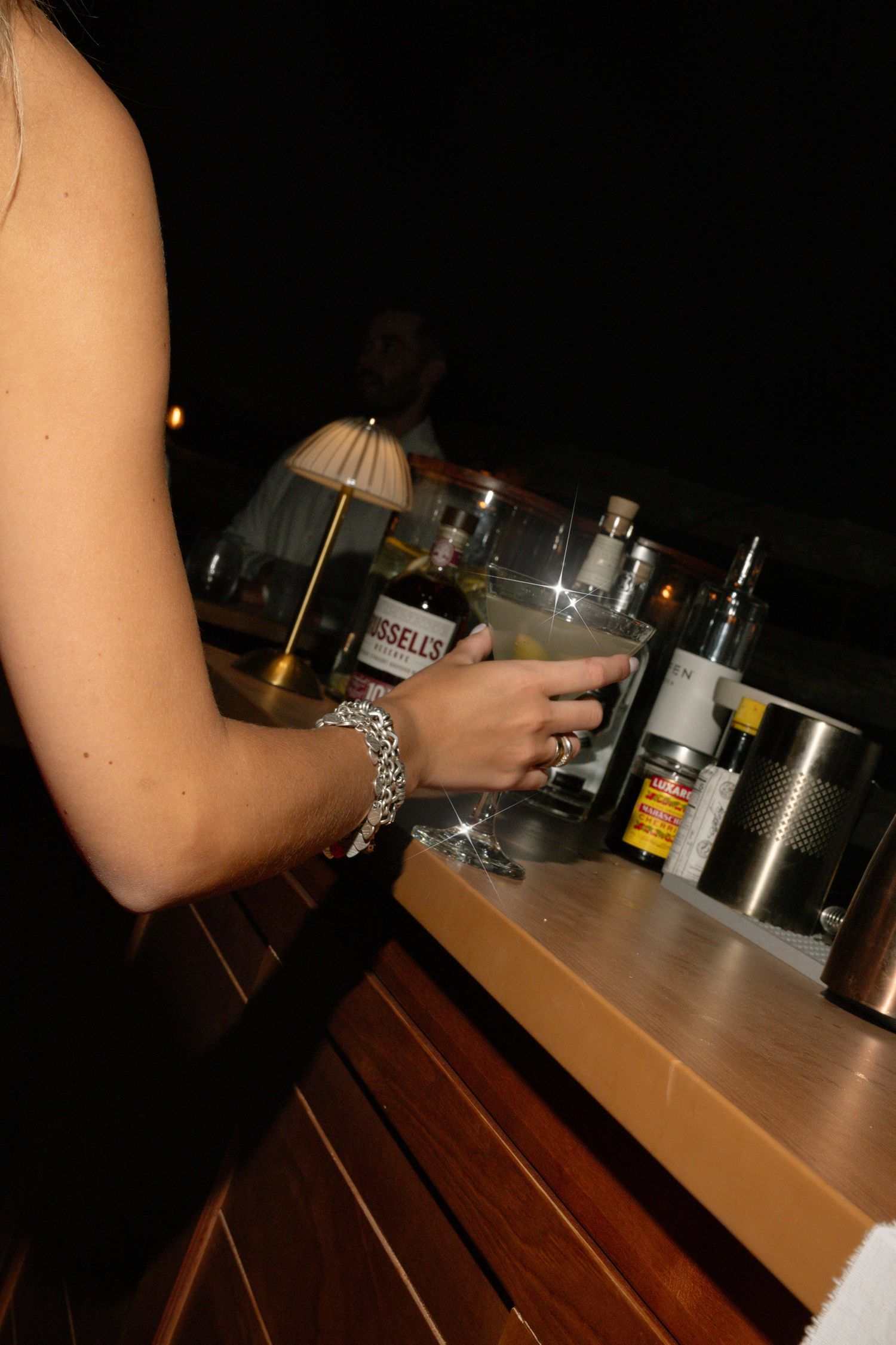 Guest holding cocktail glass at wedding bar with Russell's whiskey and bar accessories at night event.