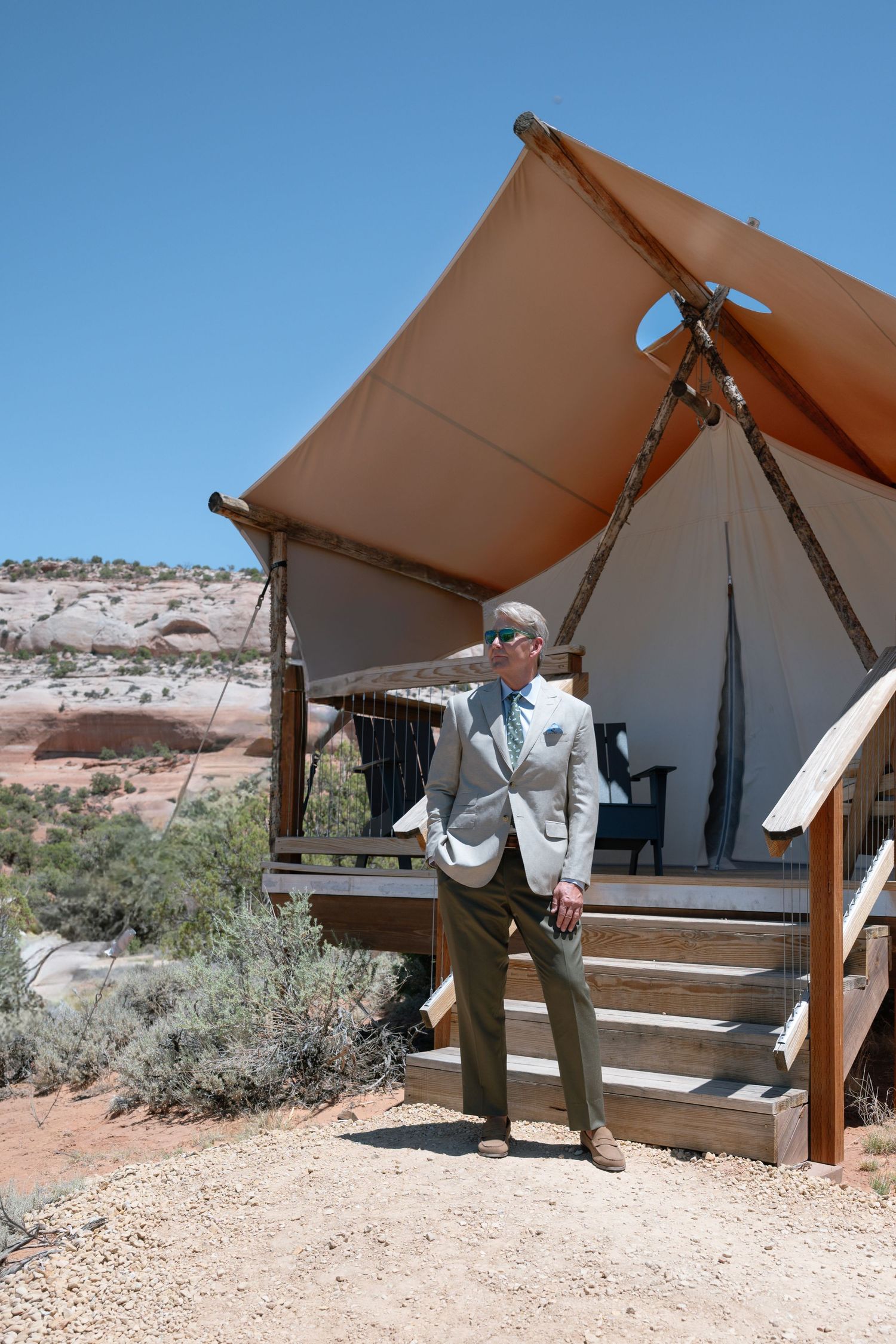 Dapper groom in tan suit stands beside a luxury safari glamping tent in red rock desert.
