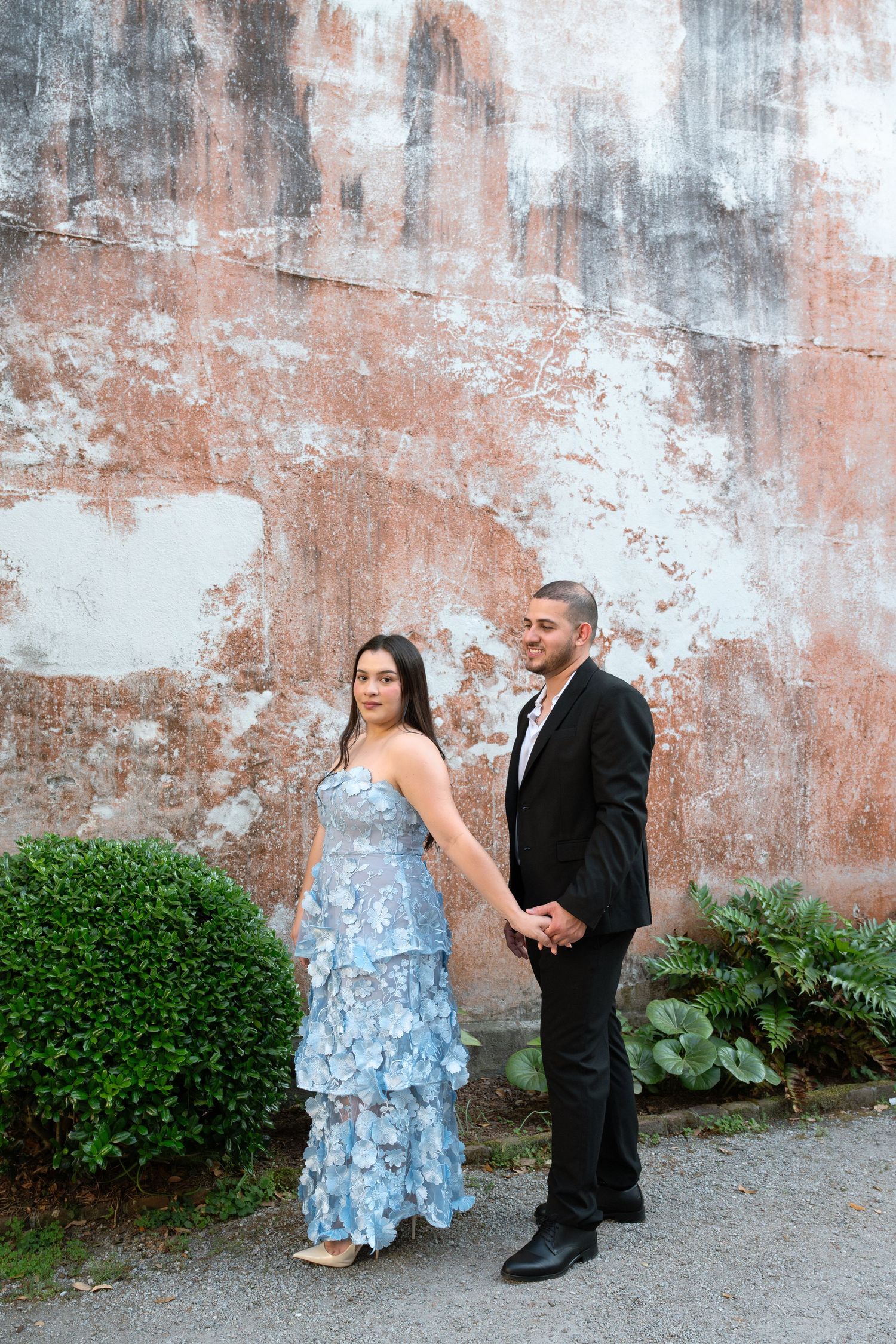 Engaged couple holding hands in blue floral dress and black suit before rustic weathered wall.