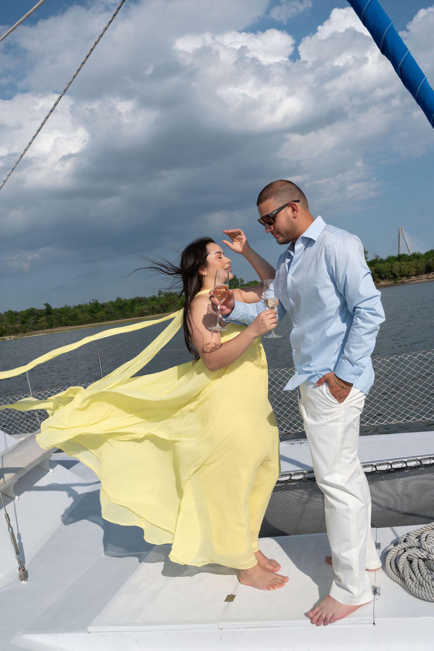 Couple enjoying champagne on a white sailboat, woman in yellow dress and man in blue shirt, with cloudy sky backdrop.