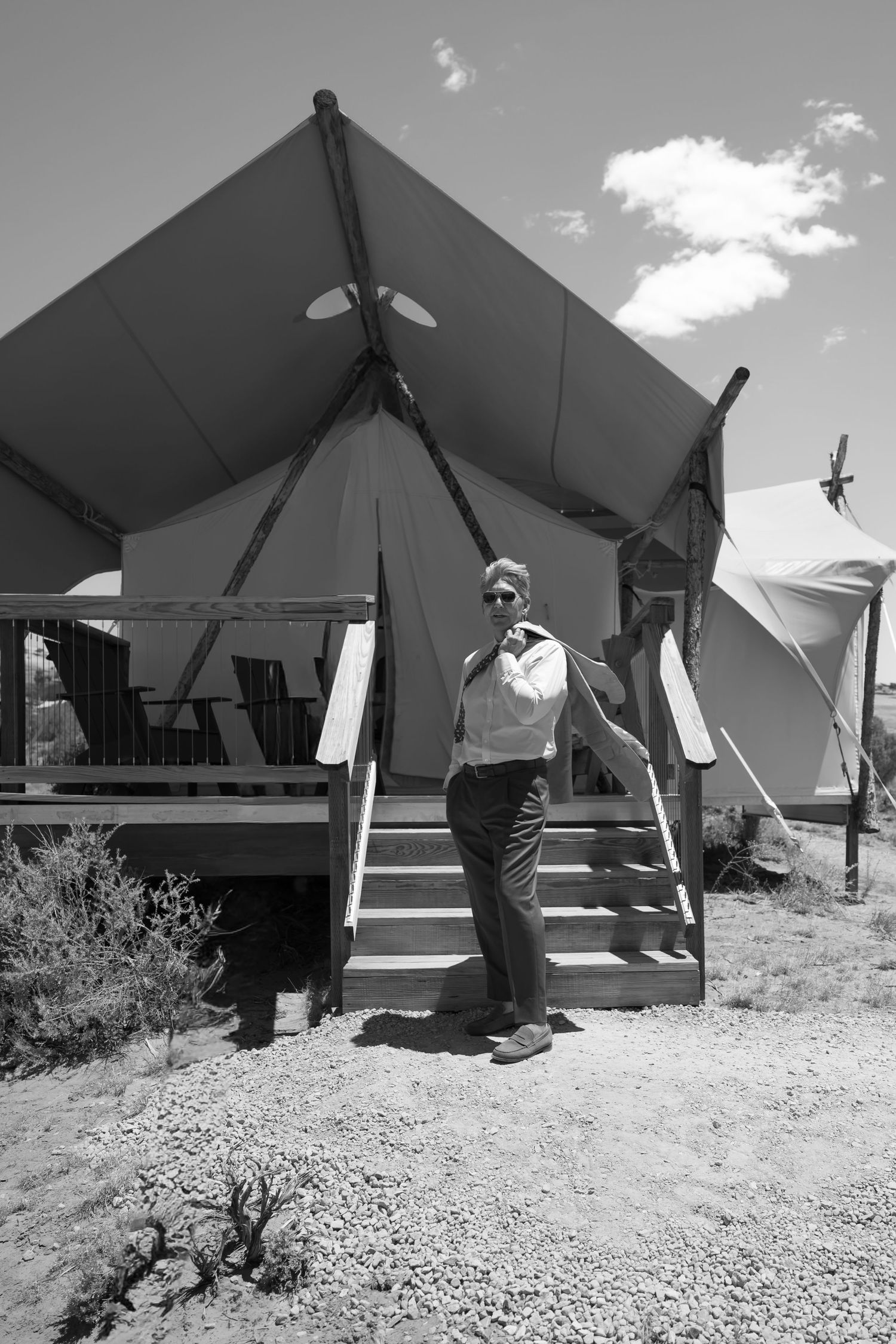 Man in dress shirt stands before a luxury canvas glamping tent in black and white desert photo.