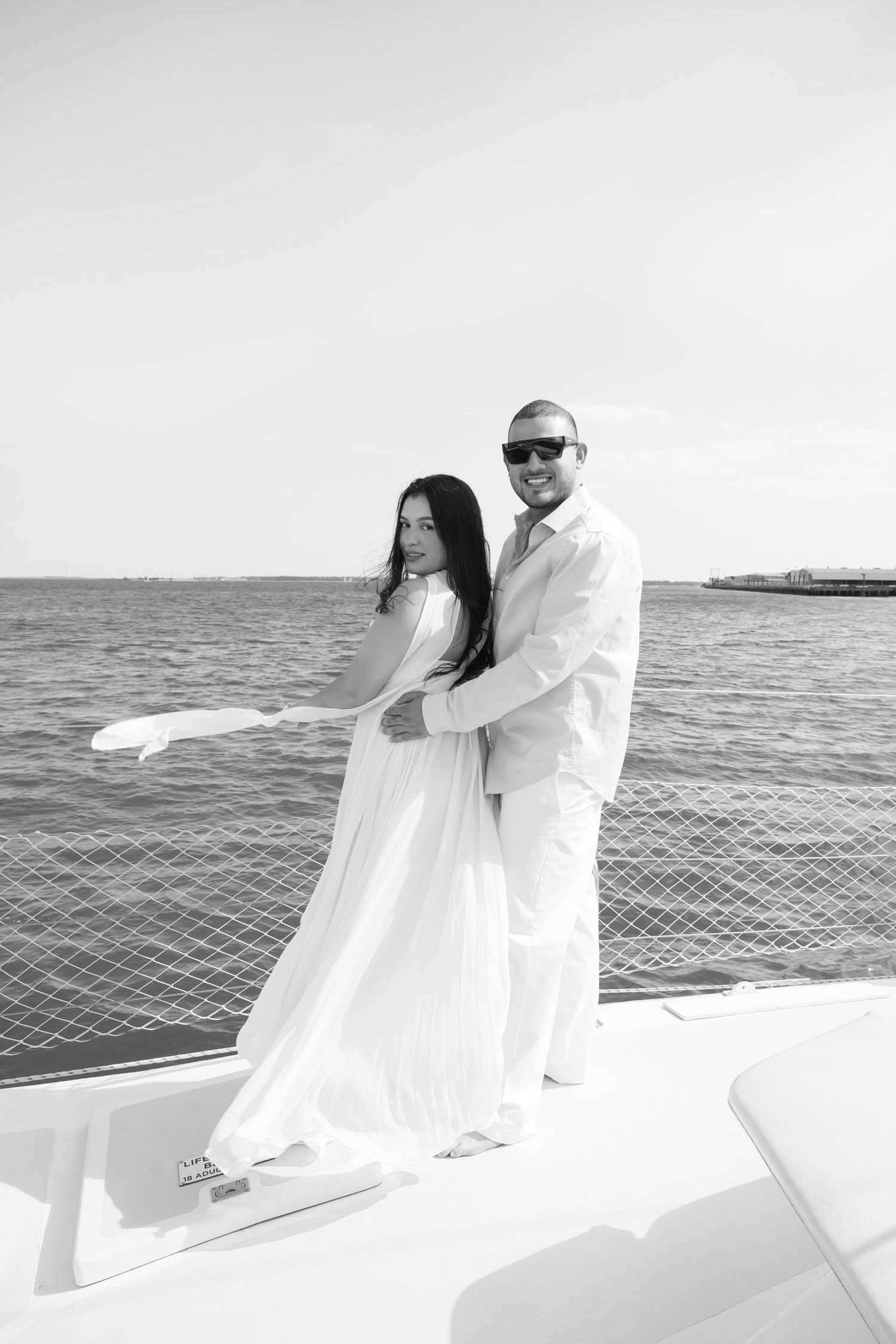 Couple in white attire embracing on a yacht bow with ocean backdrop in black and white photo.