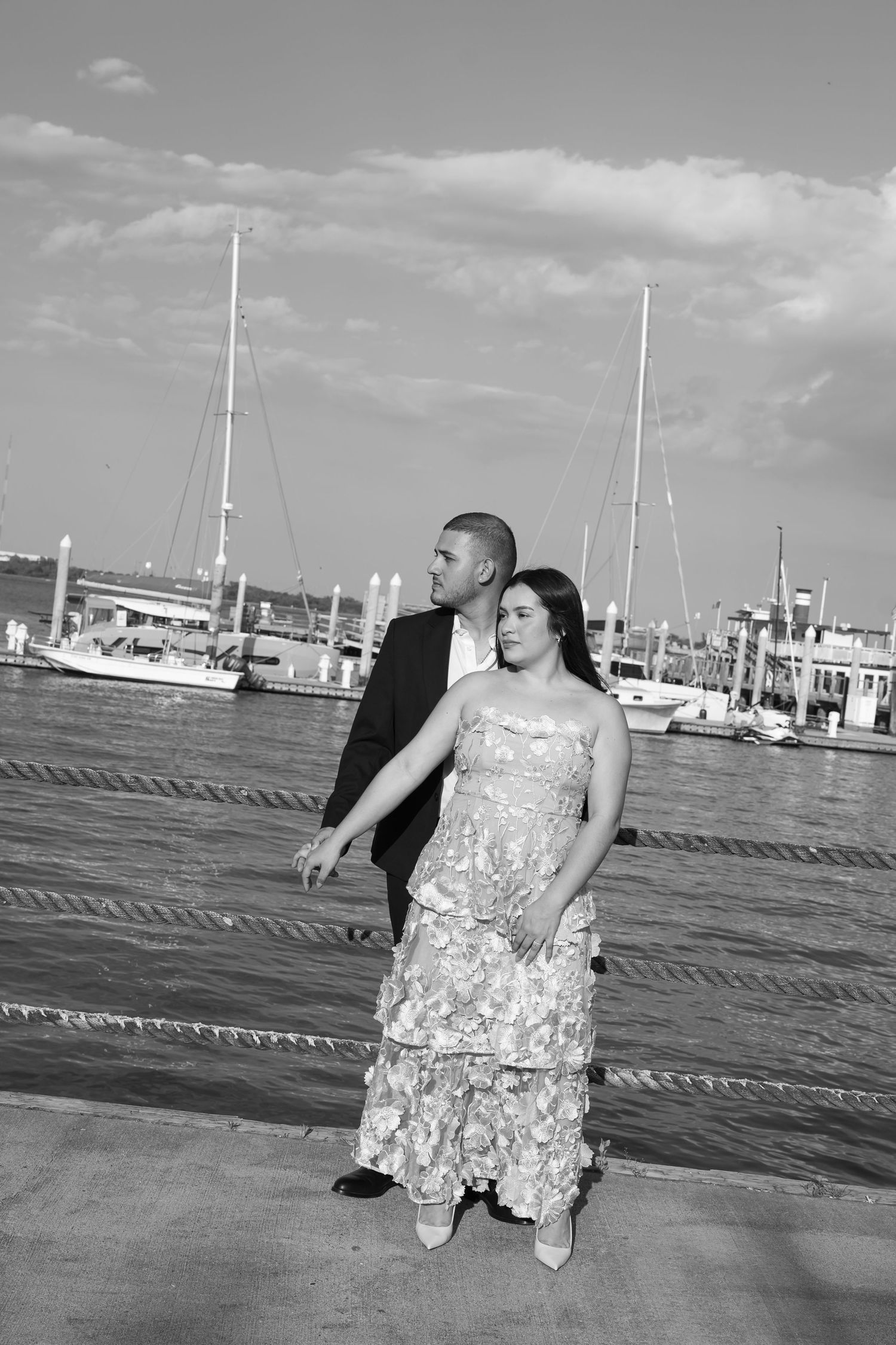 Couple posing on a marina dock, man in suit embracing woman in floral gown, sailboats in background.