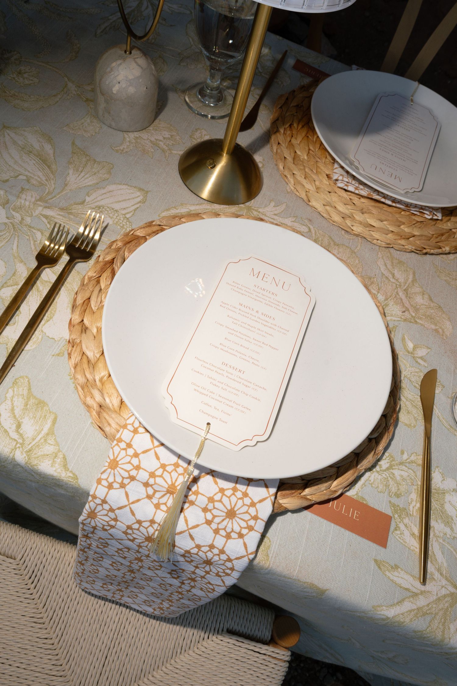 Elegant wedding place setting with woven charger, white plate, menu card, gold cutlery, and patterned napkin.
