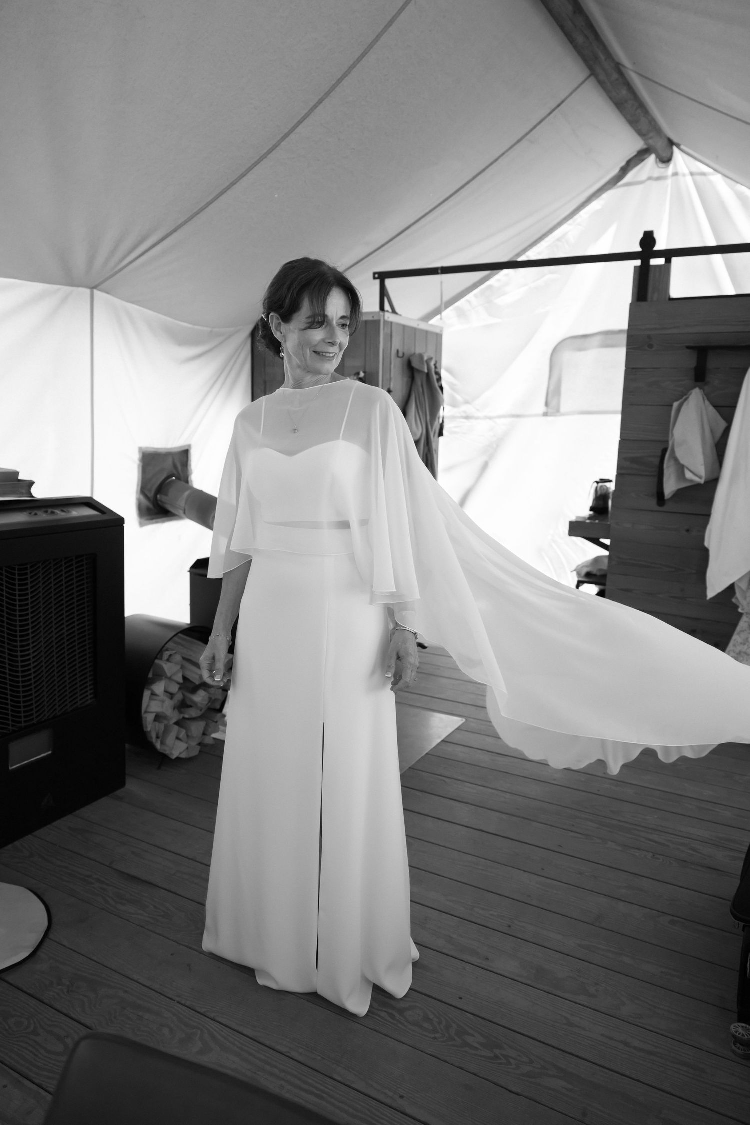 Bride in elegant white flowing cape wedding gown smiling inside a tent venue in black and white photo.