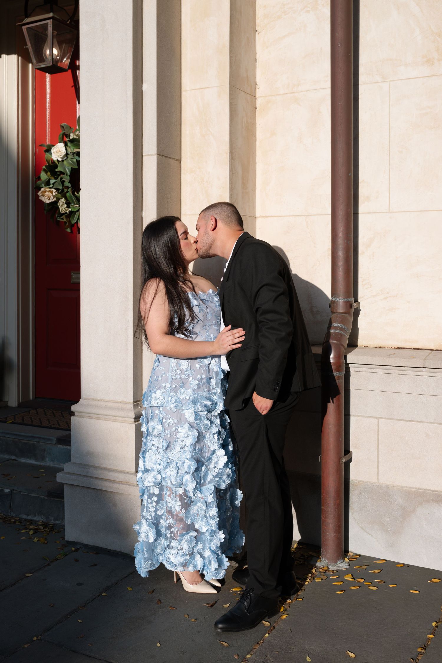 Couple sharing a romantic kiss outdoors, woman in blue floral gown and man in black suit beside a column.