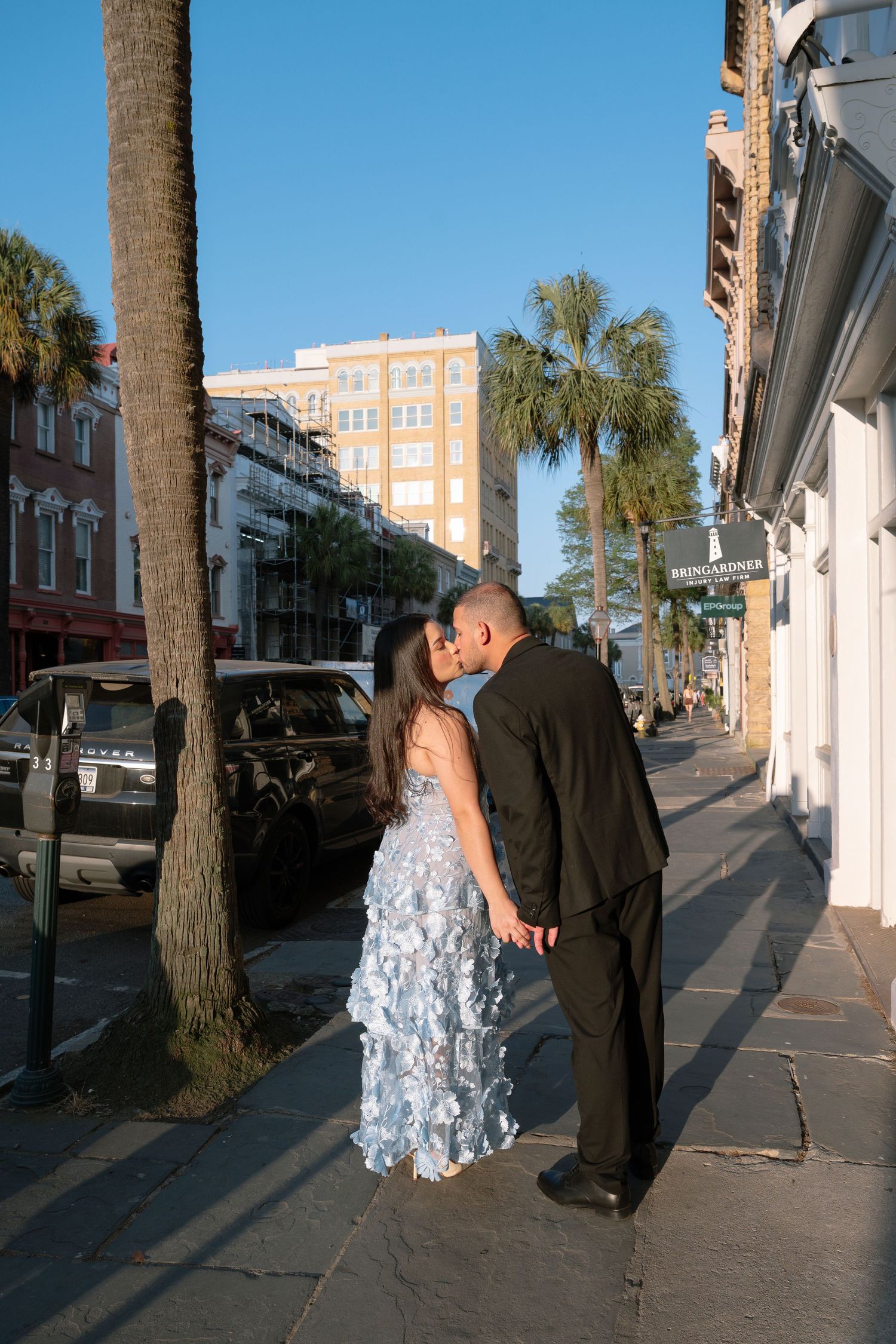 Couple sharing a romantic kiss on a palm-lined Charleston street, dressed in elegant formal attire.
