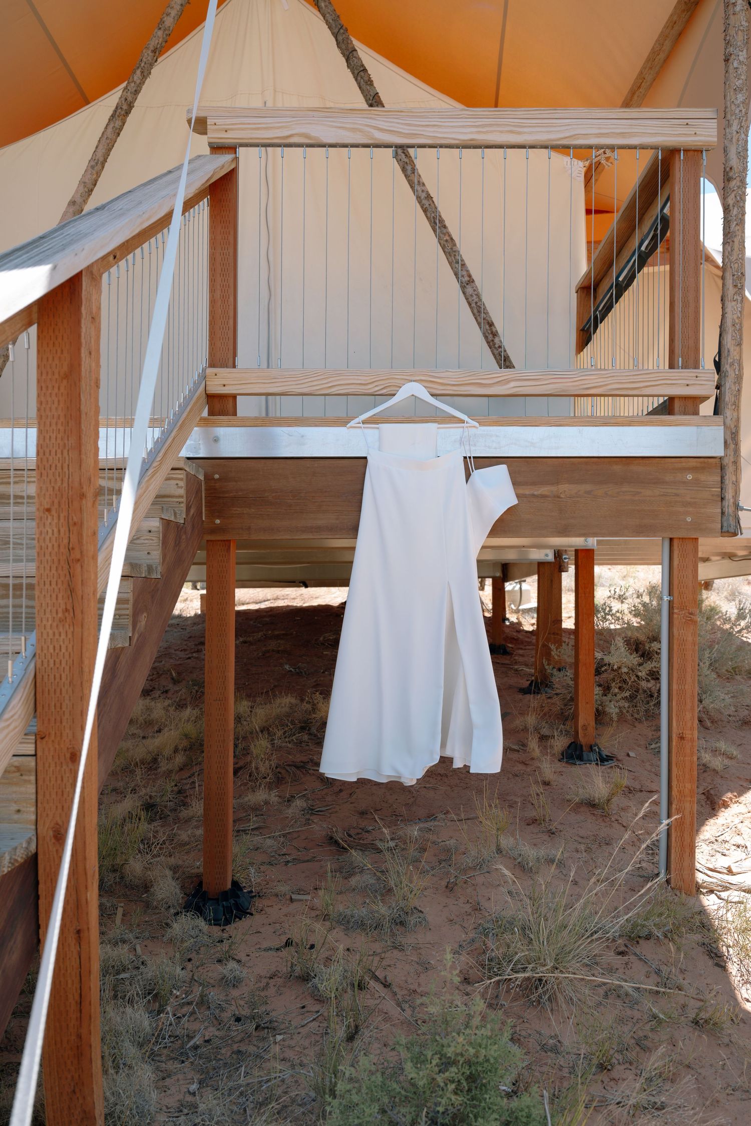 White wedding dress hanging on wooden deck of desert glamping tent structure.