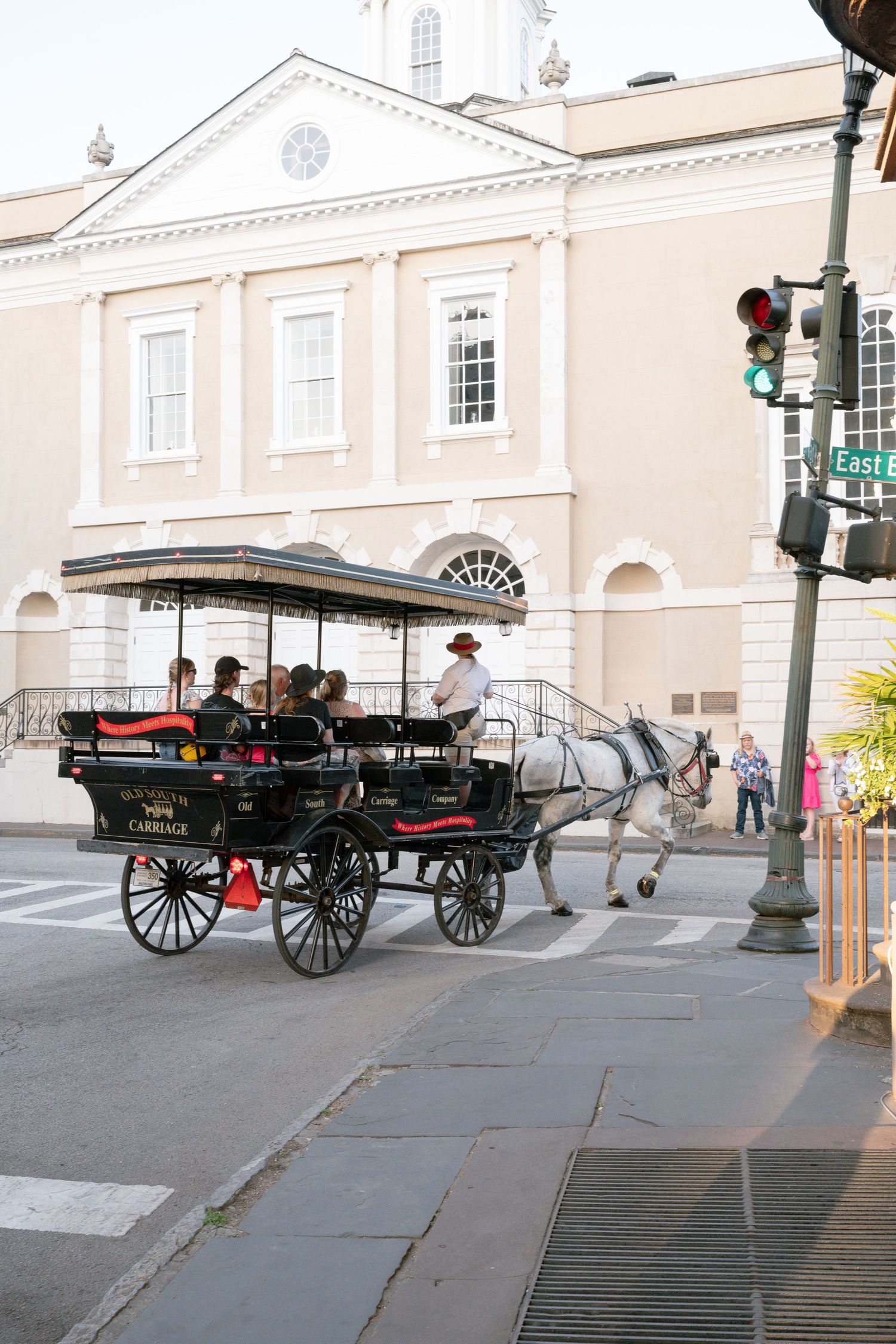 Historic horse-drawn carriage tour passing a neoclassical building at a sunny intersection in Charleston, SC.