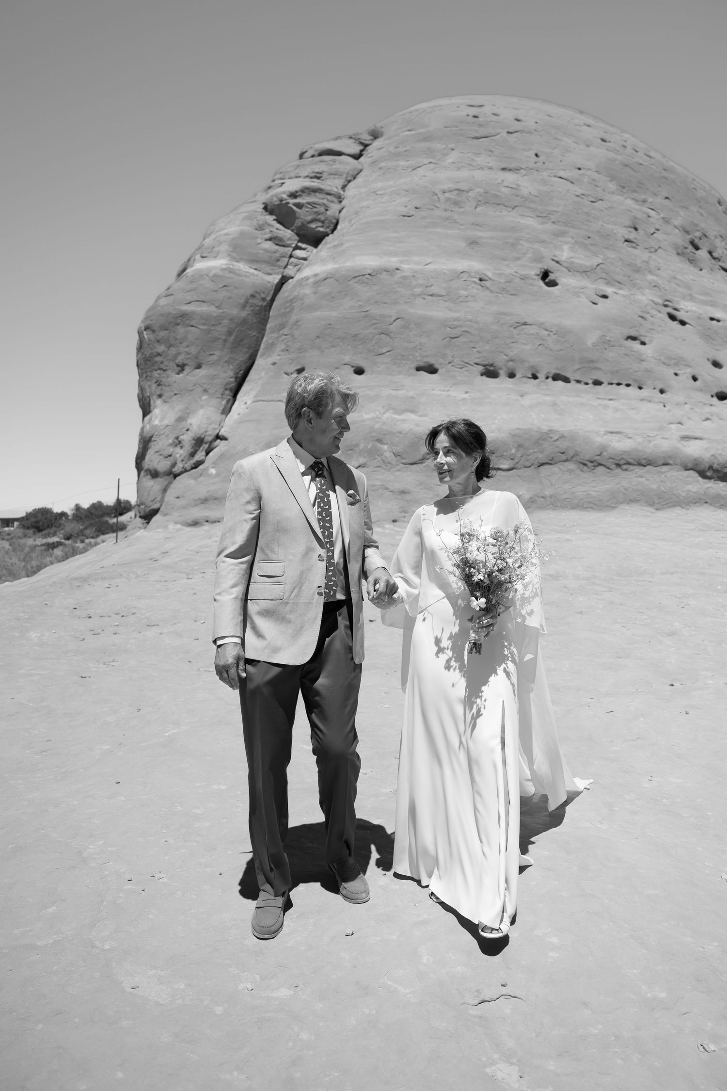 Black and white photo of bride and groom walking together near red rock formation in desert landscape.