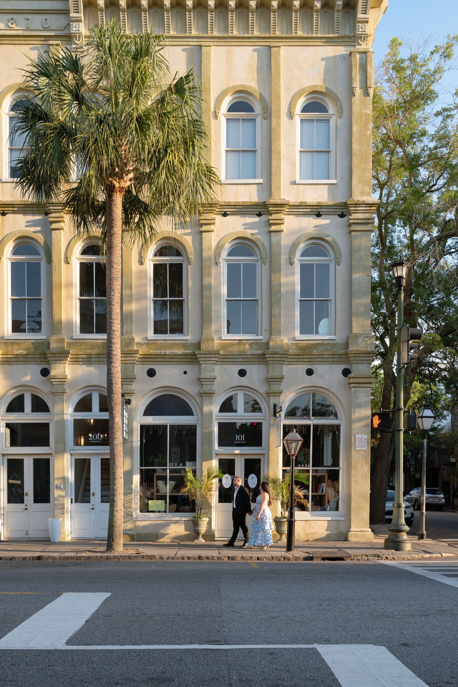 Bride and groom walking past a historic yellow building with arched windows and a palm tree in Charleston, SC.