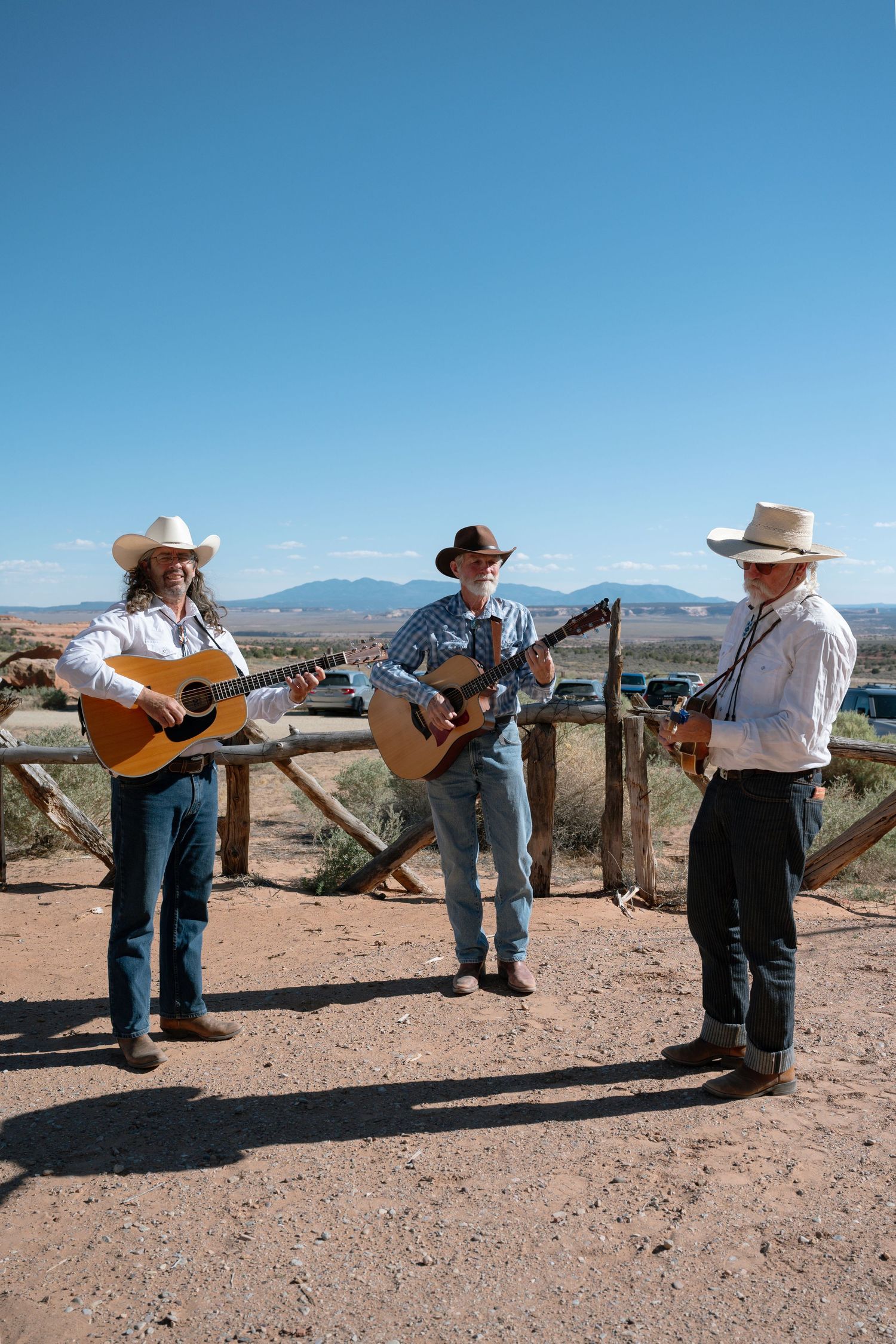 Three cowboys in white shirts and hats playing acoustic guitars and fiddle outdoors in a desert landscape.