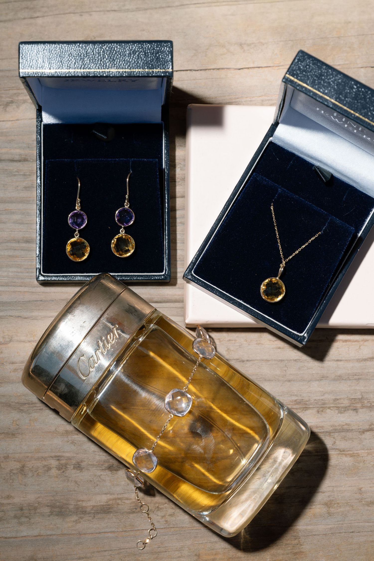 Cartier perfume bottle beside two open jewelry boxes displaying gold earrings with gemstones and a delicate pendant necklace.