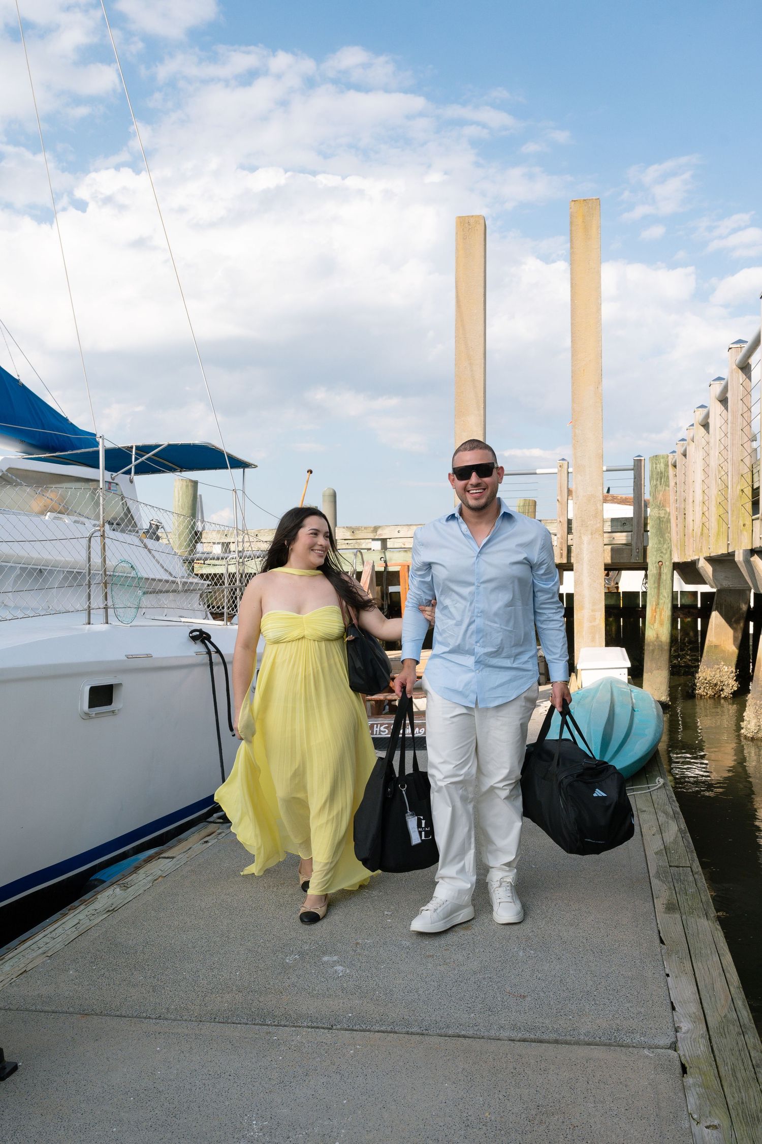 Couple walking along marina dock carrying bags, woman in yellow maxi dress, man in light blue shirt and white pants.