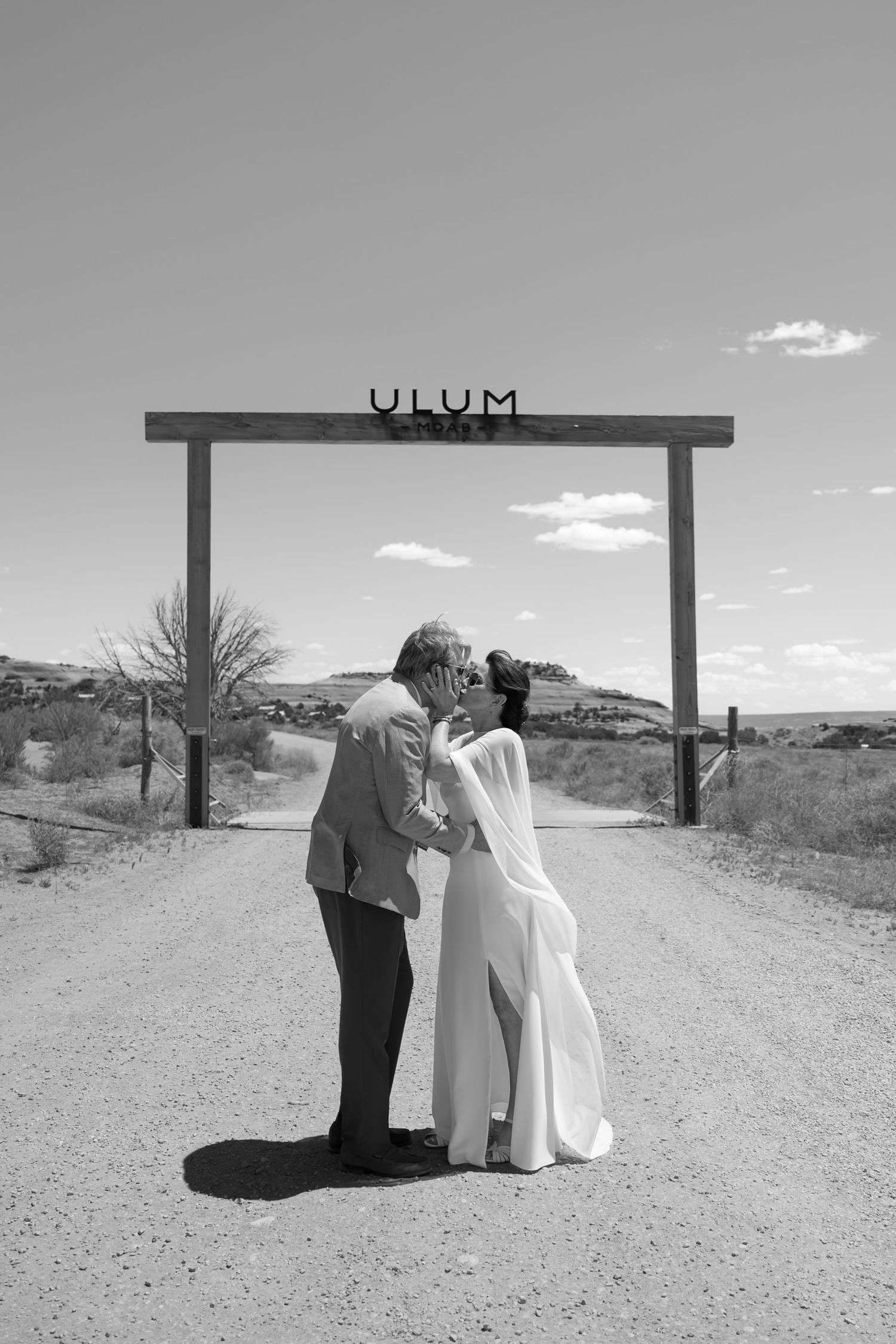 Black and white photo of couple kissing under rustic ULUM sign on a desert dirt road.