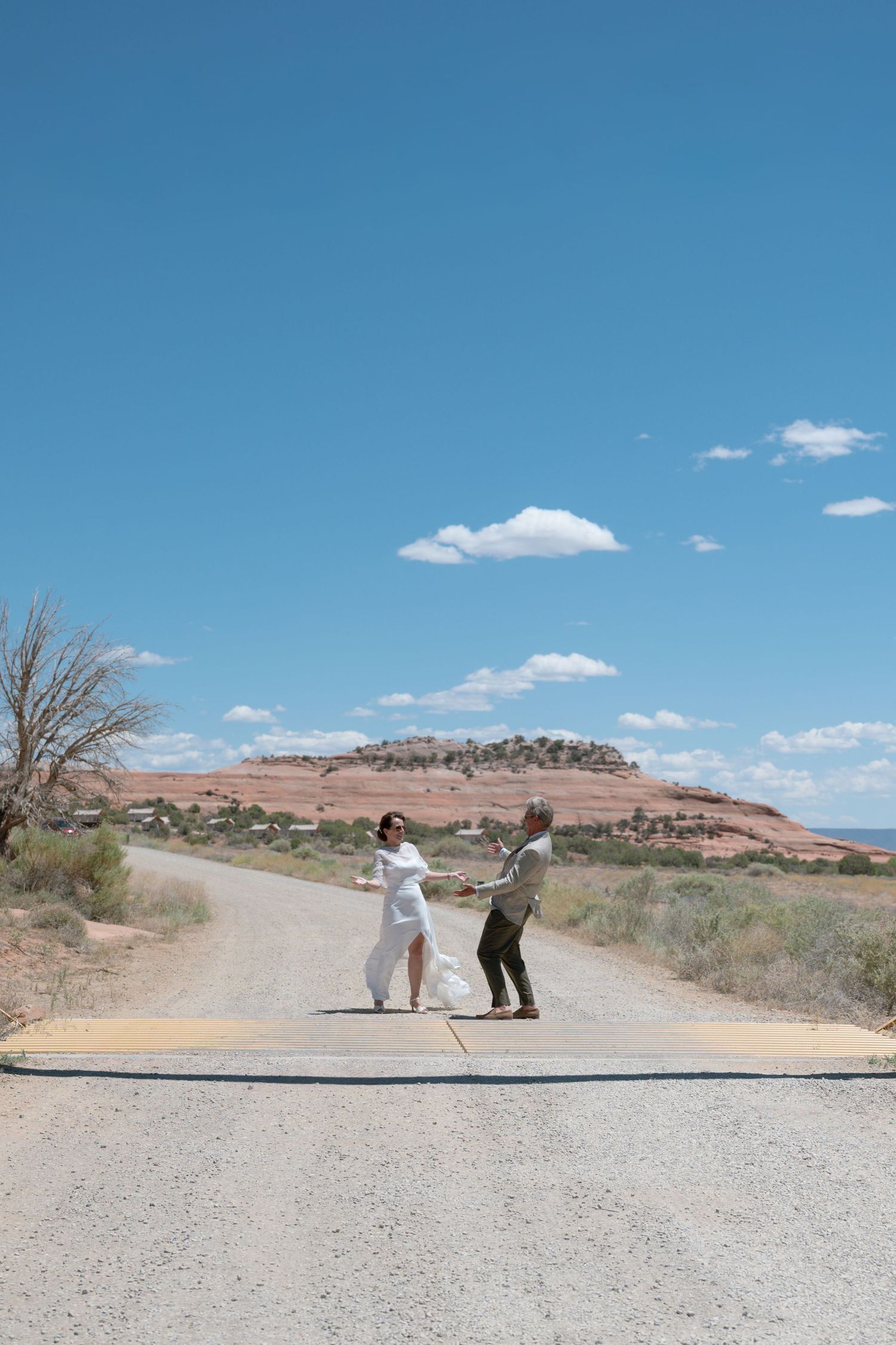 Bride and groom dance joyfully on a remote desert road with red rock mesa and blue sky backdrop.