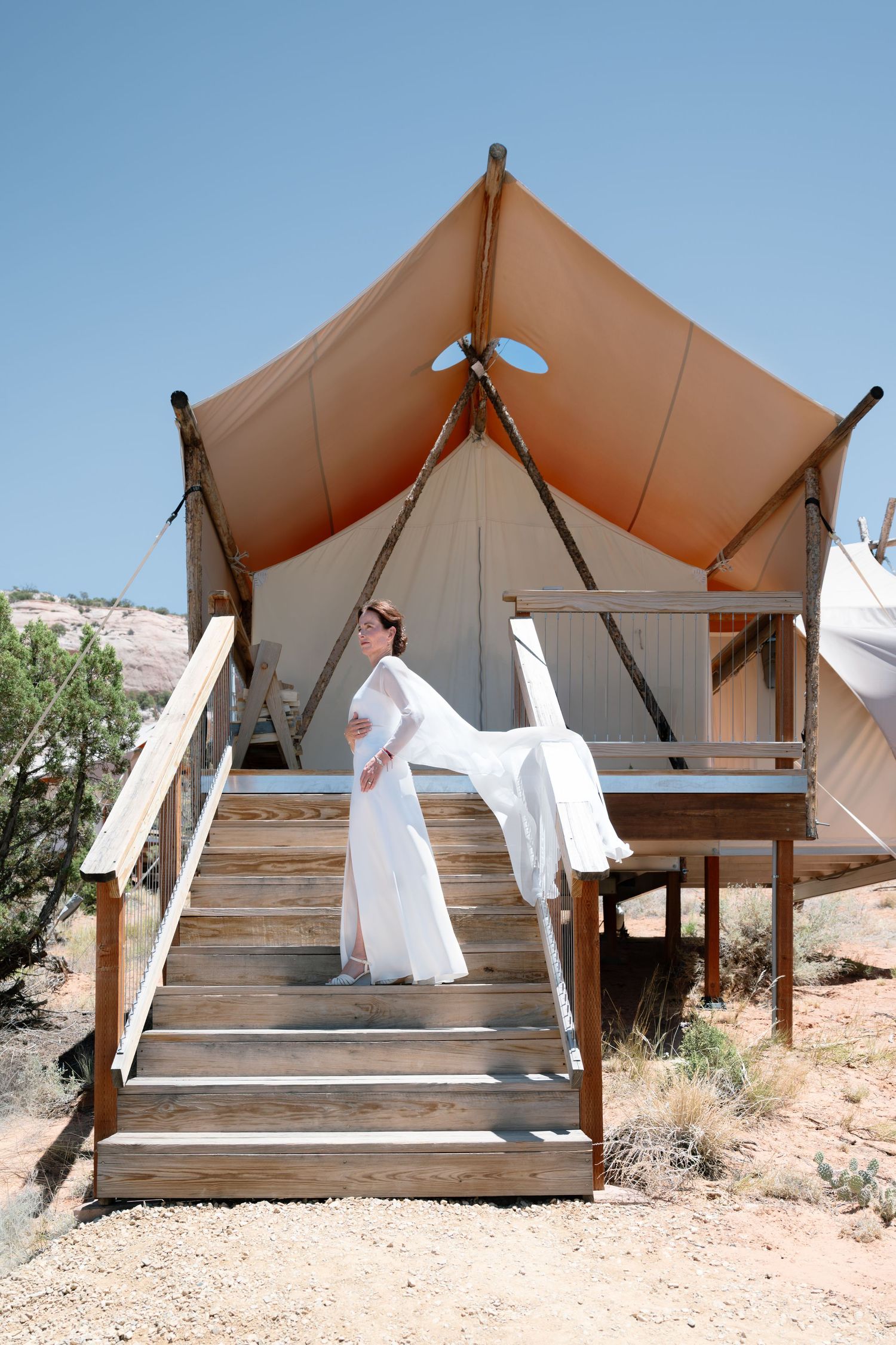 Bride in flowing white gown stands on wooden stairs of a glamping tent in arid desert landscape.