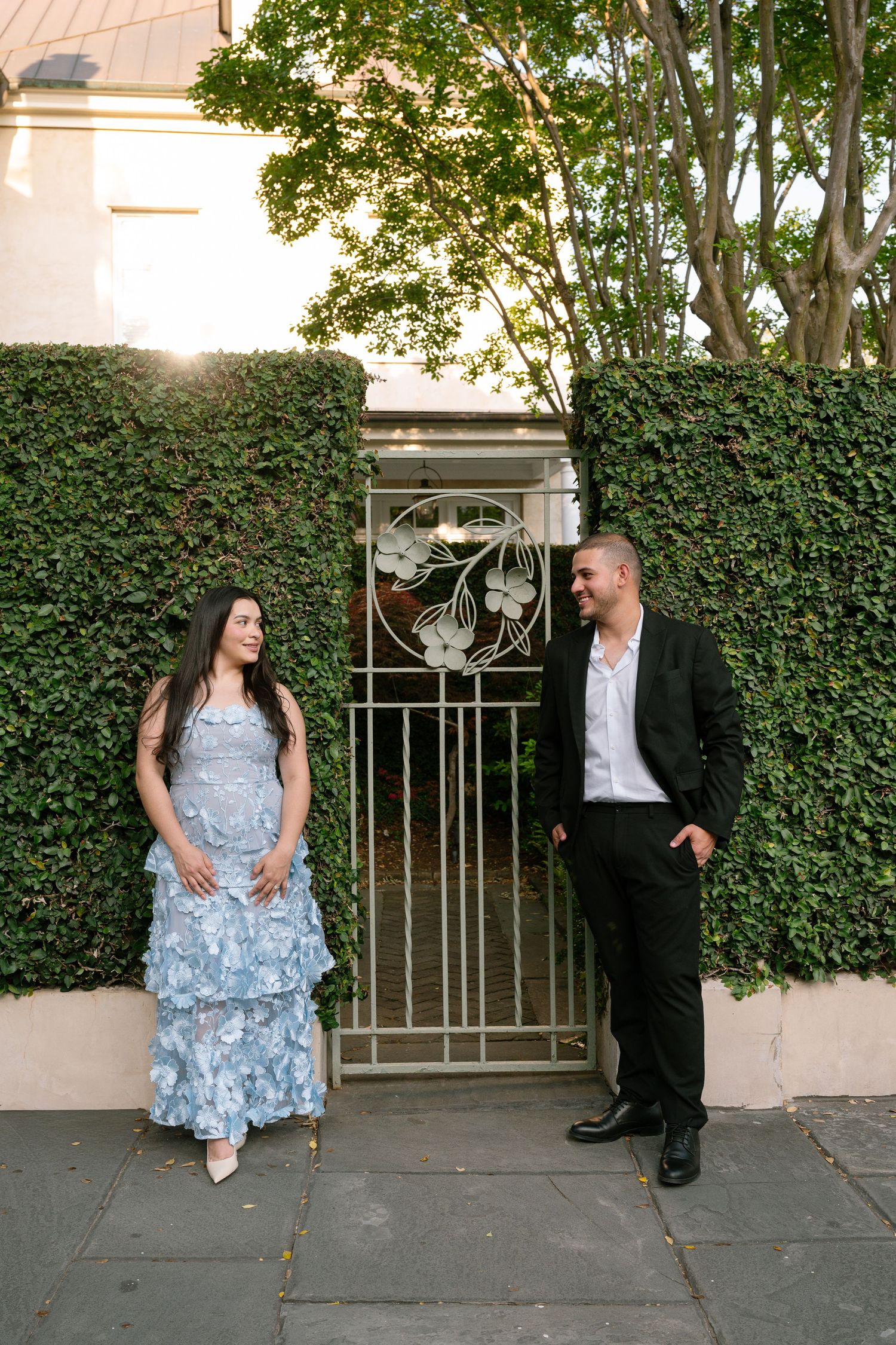 A couple stands by an ornate iron gate covered in ivy, woman in blue floral gown, man in black suit, smiling at each other.