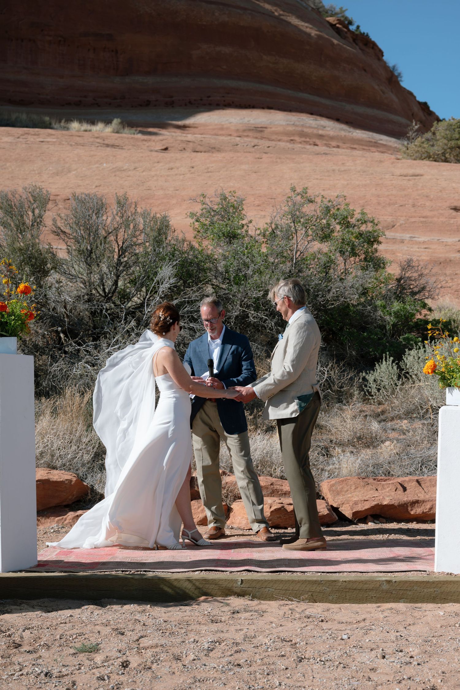 Outdoor desert wedding ceremony with bride and groom exchanging vows before an officiant, red rock formations in background.