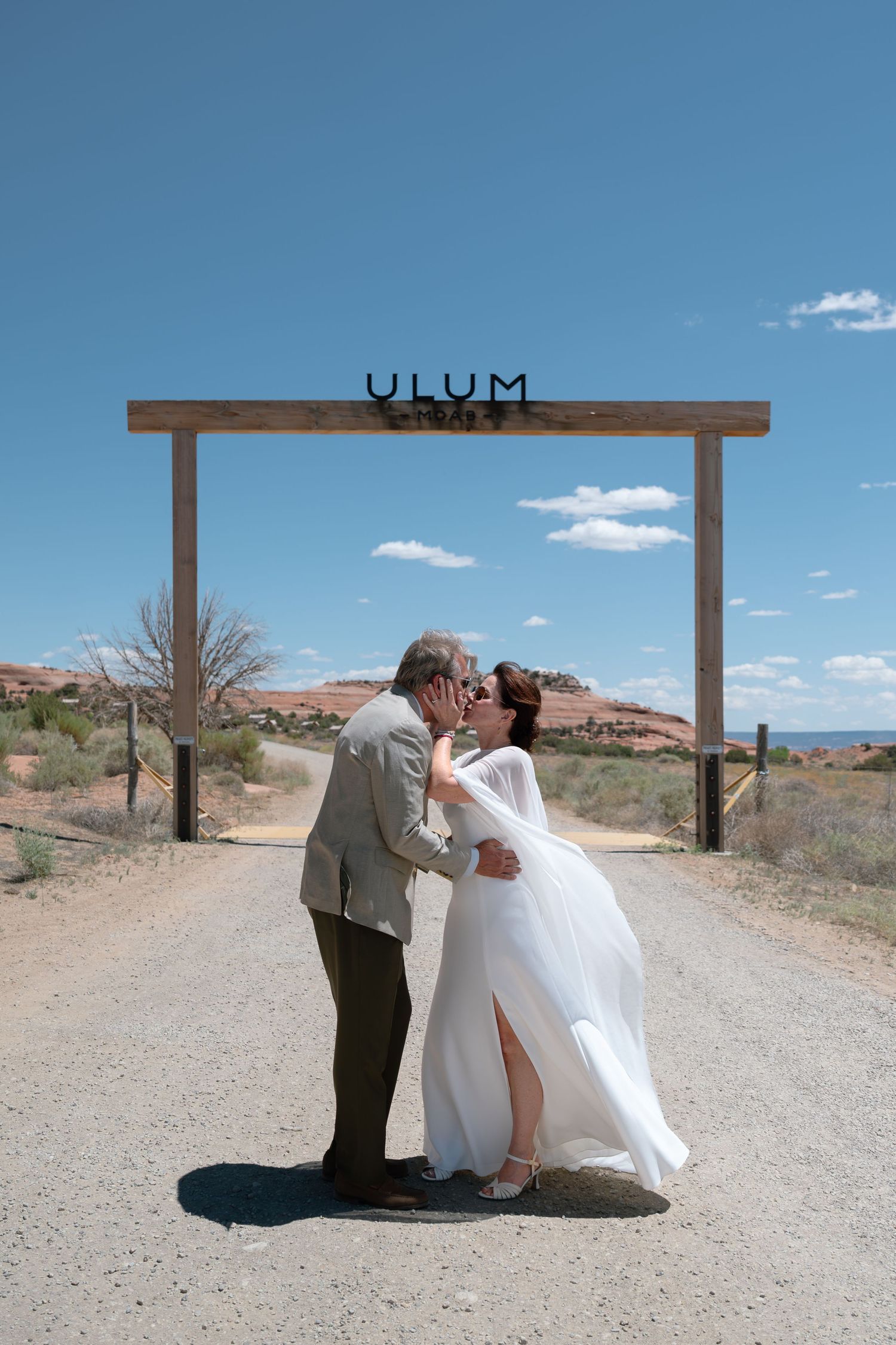 Couple sharing a first kiss under a rustic wooden ULUM sign on a desert dirt road on their wedding day.