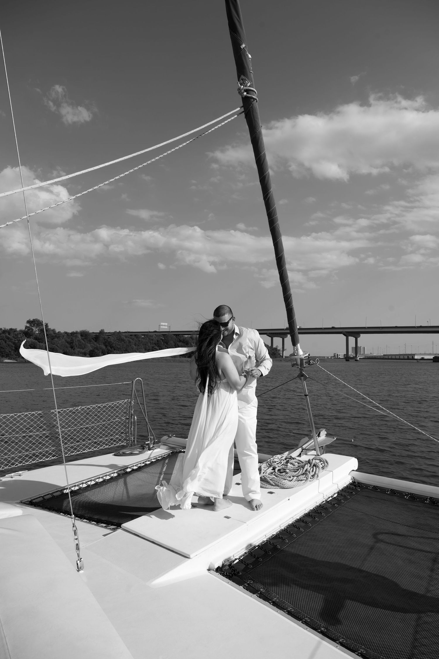 Black and white photo of a couple embracing on a catamaran sailboat deck with a bridge and calm water in the background.