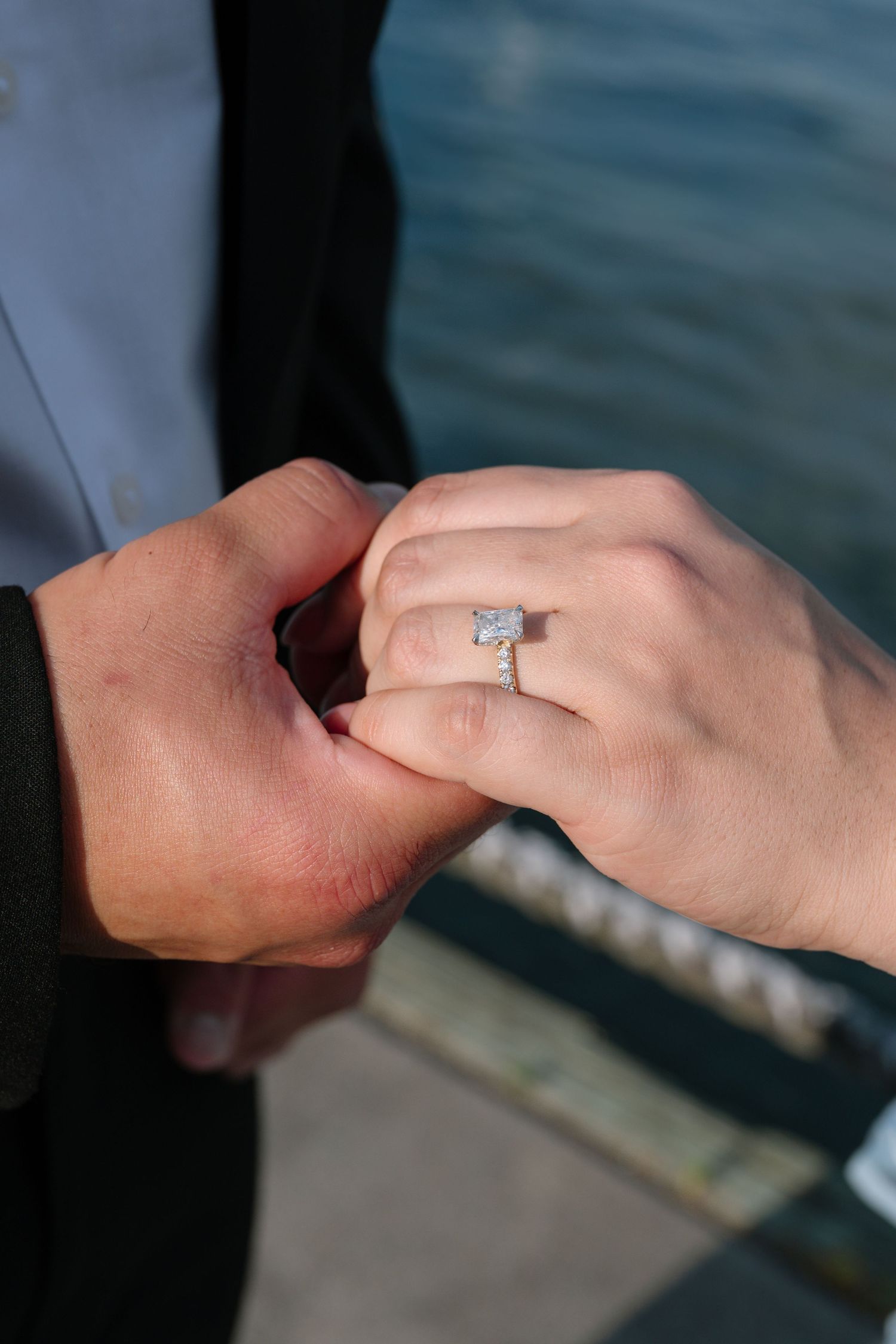 Close-up of engagement ring with emerald cut diamond on woman's hand held by partner near waterfront.