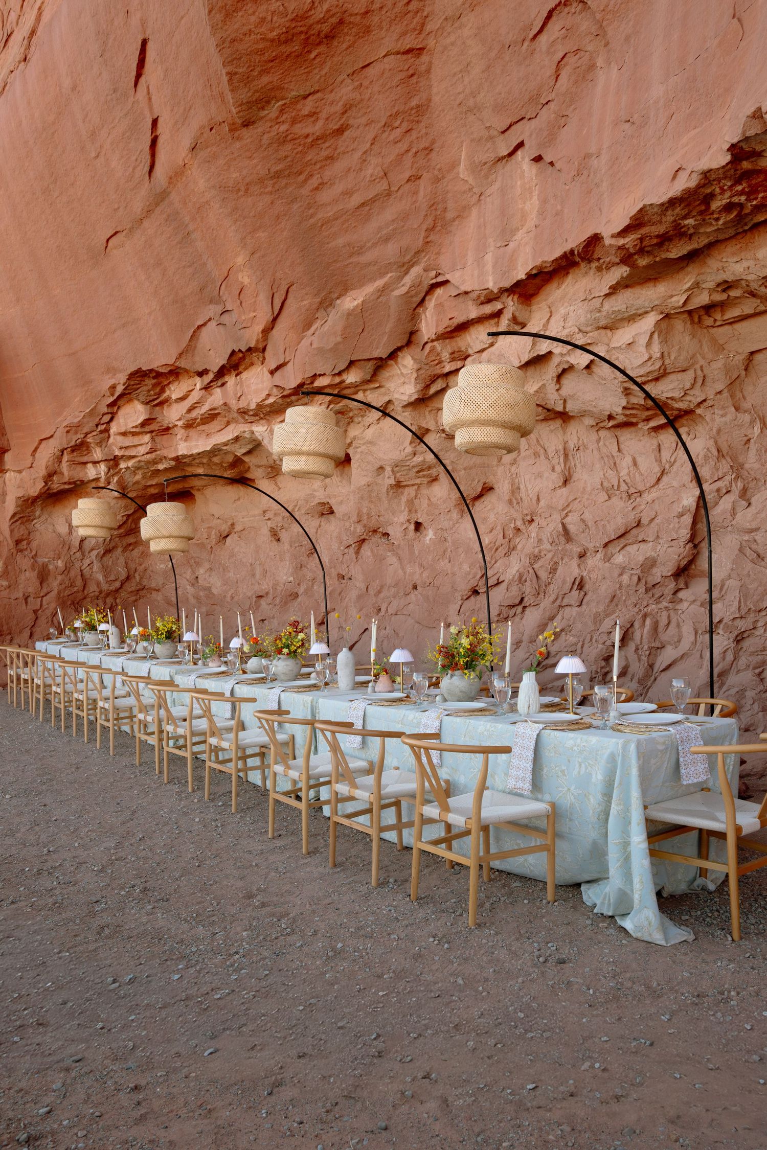 Elegant outdoor wedding reception table set beneath red canyon wall with arched rattan pendant lights.