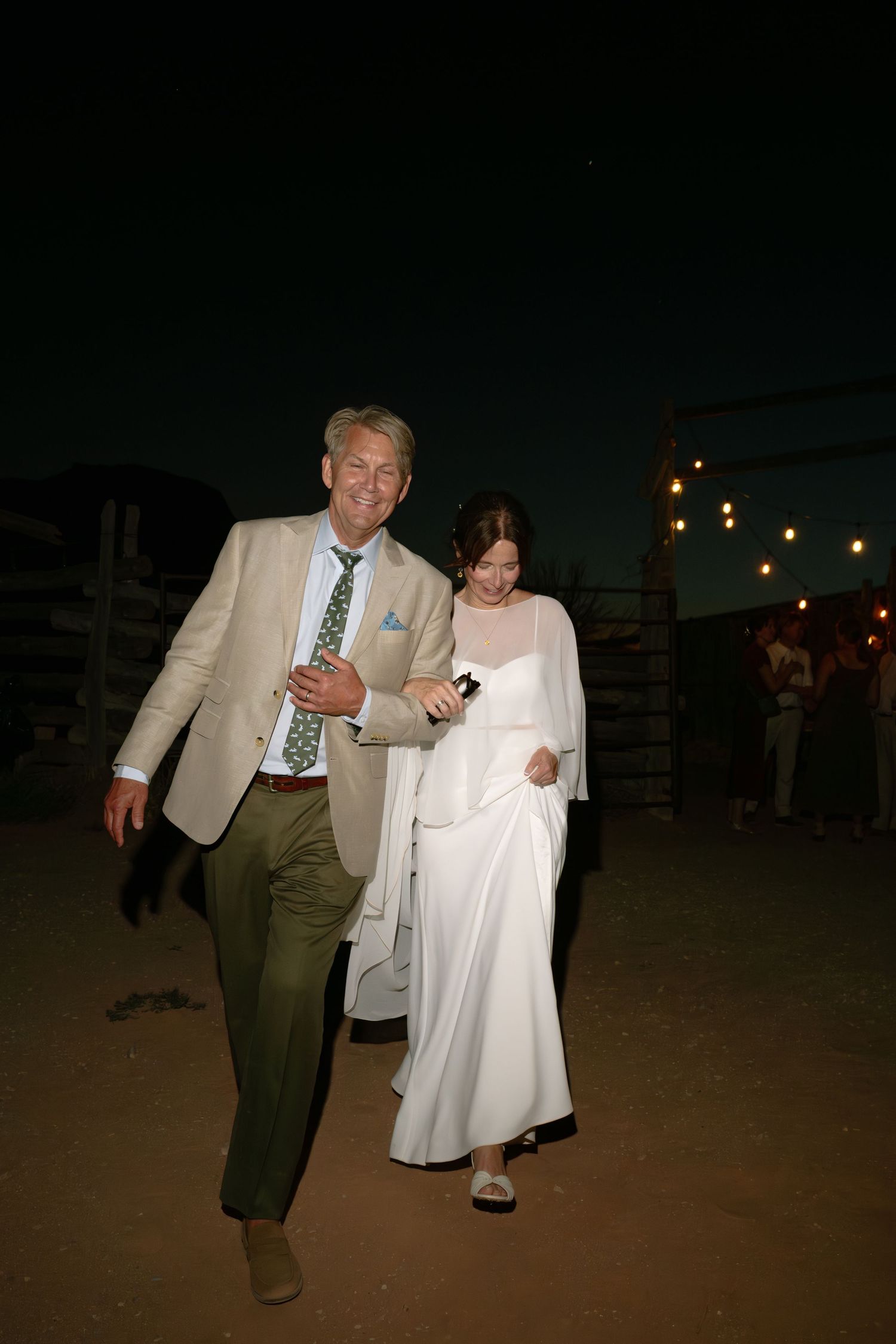 Bride in white gown and groom in tan suit with green tie walk together outdoors at night wedding reception.