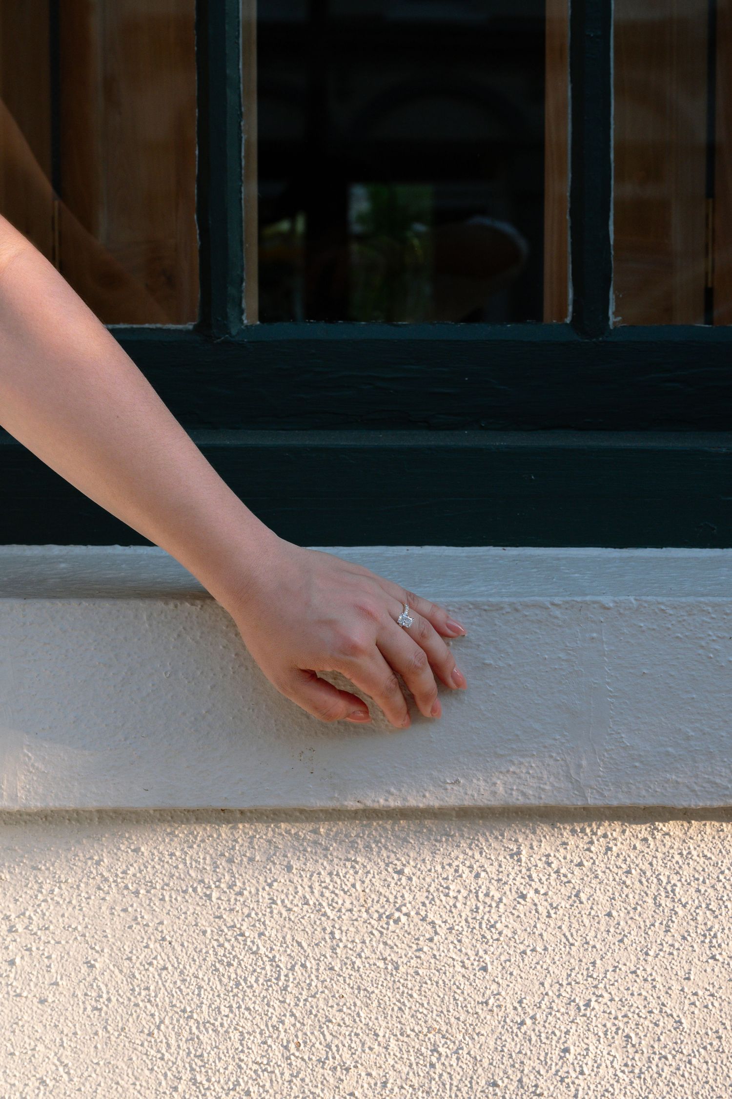 A woman's hand with a silver ring resting on a white textured wall below a dark green window frame.