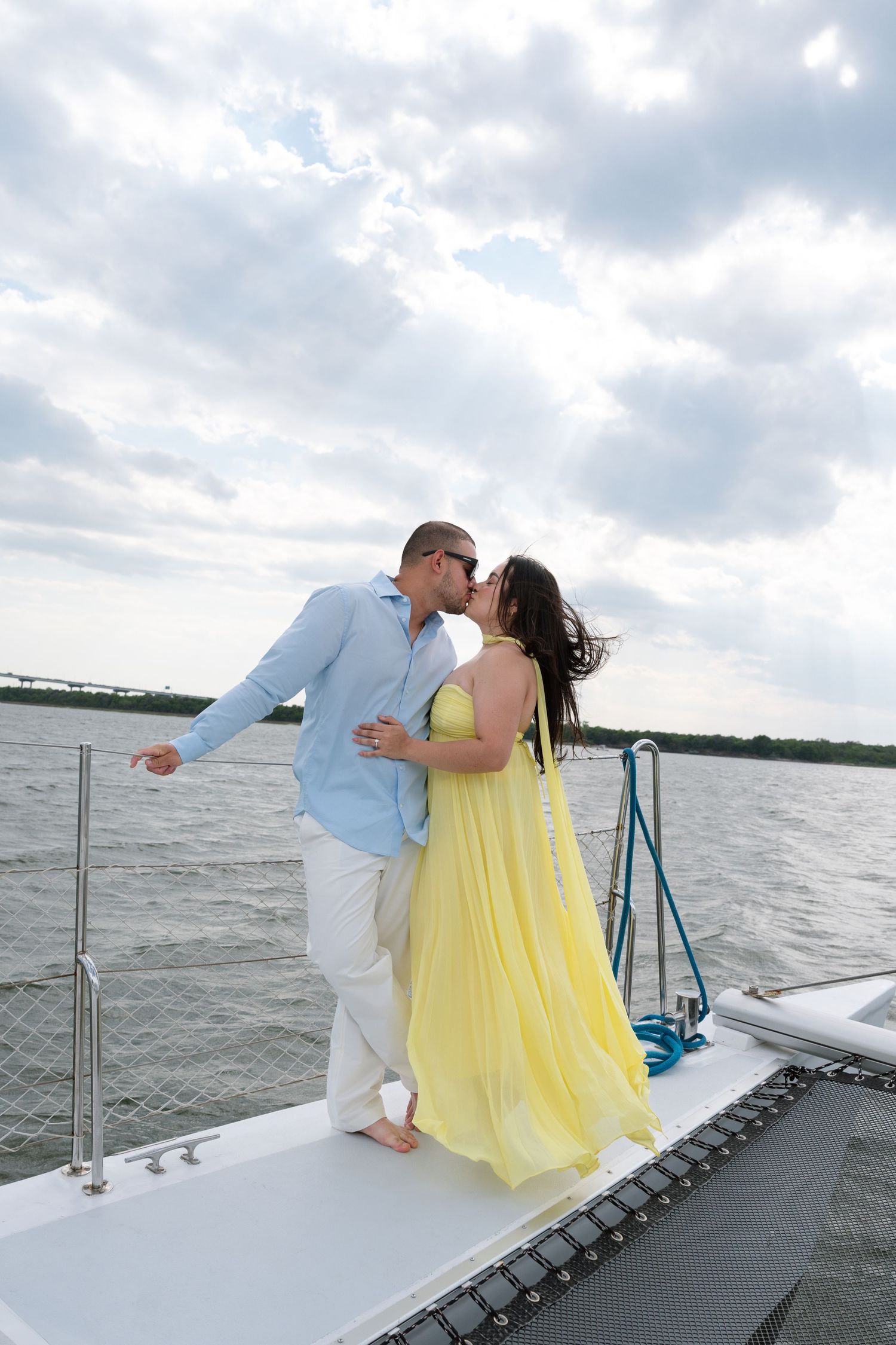 Couple sharing a romantic kiss on a white catamaran deck, woman in yellow dress, cloudy sky over water.
