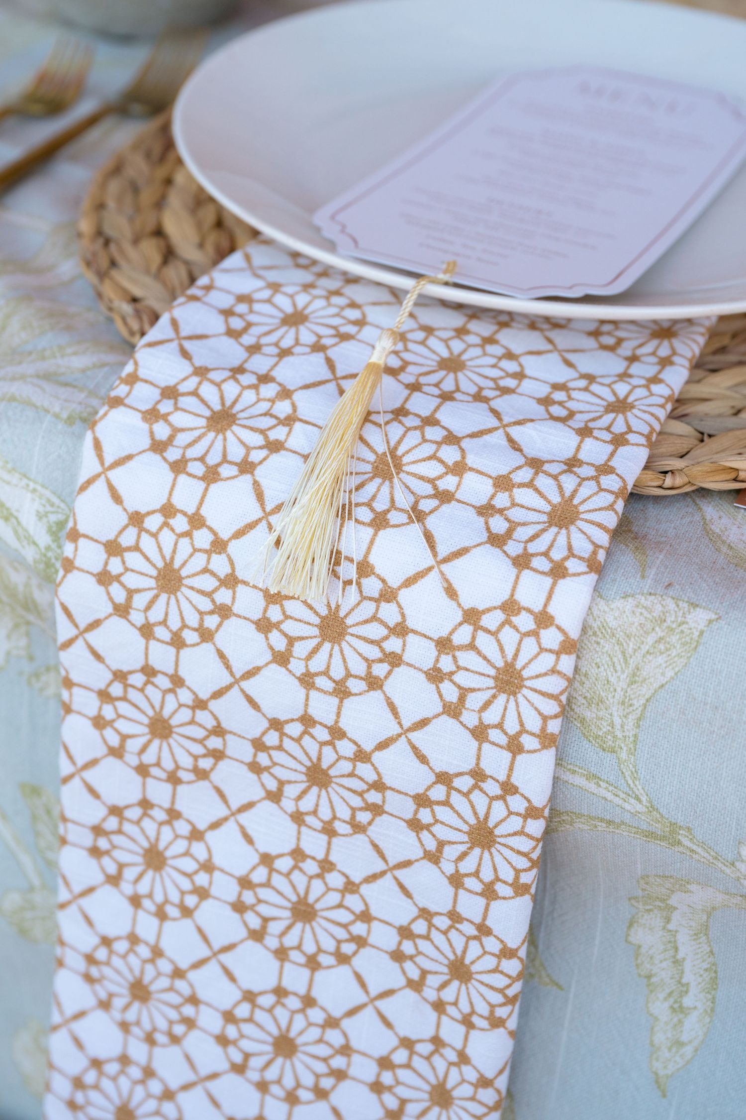 An elegant table setting featuring a gold geometric patterned napkin with tassel on a woven placemat beside a menu card.