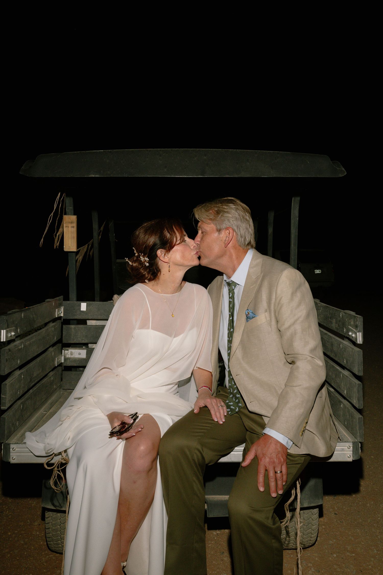 A couple shares a romantic kiss while seated on the back of a rustic vehicle at an outdoor nighttime wedding celebration.