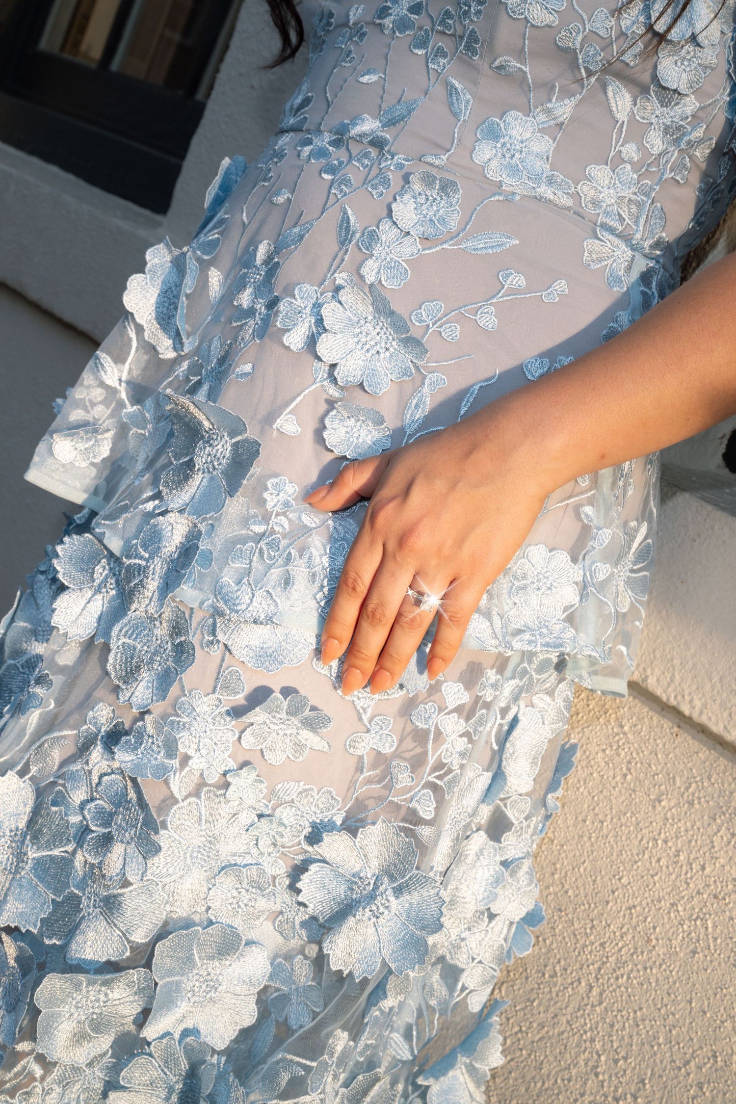 Close-up of an engagement ring on a woman's hand resting on a blue floral embroidered lace dress.