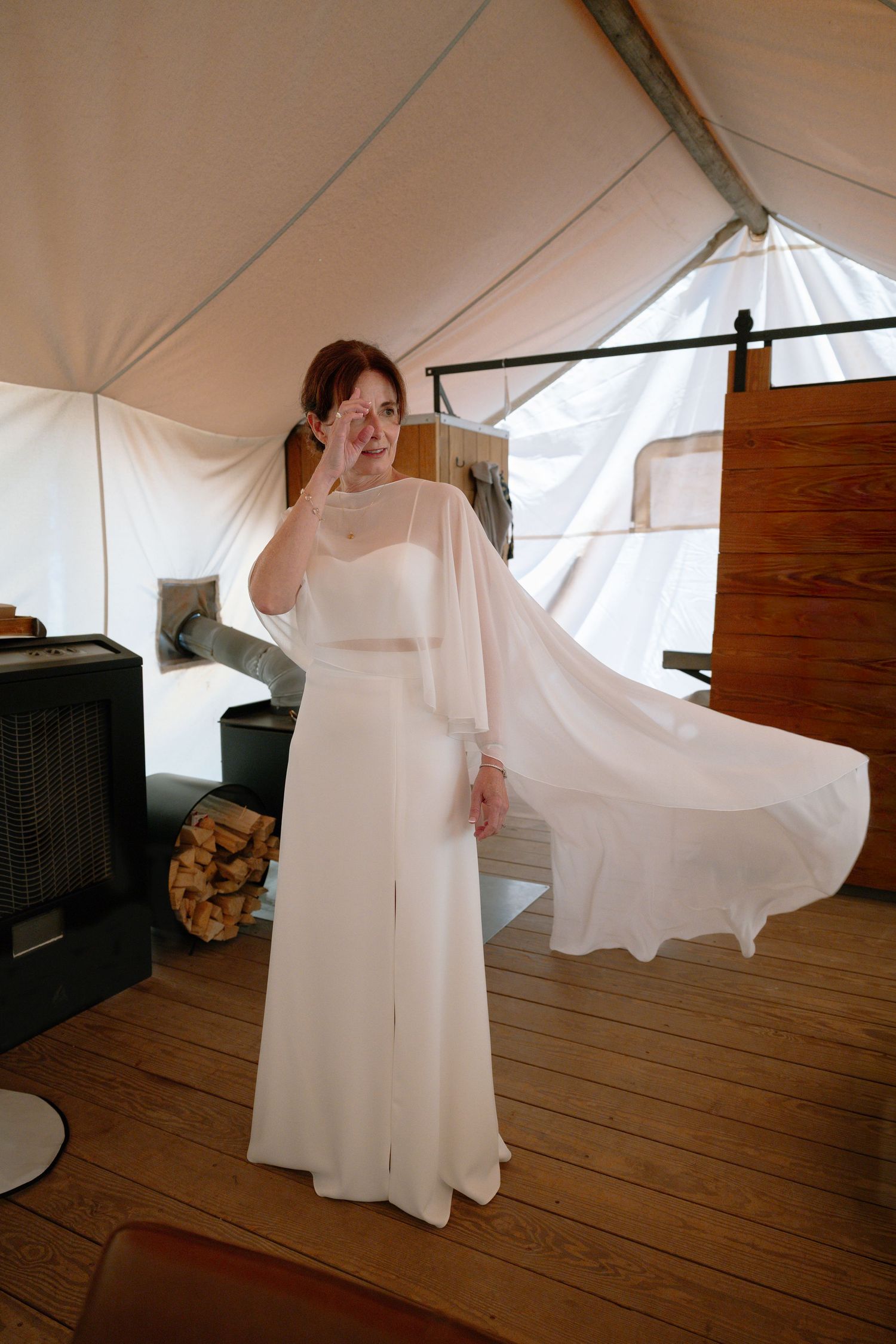 Bride in ethereal white cape wedding gown poses inside a luxurious glamping tent interior.