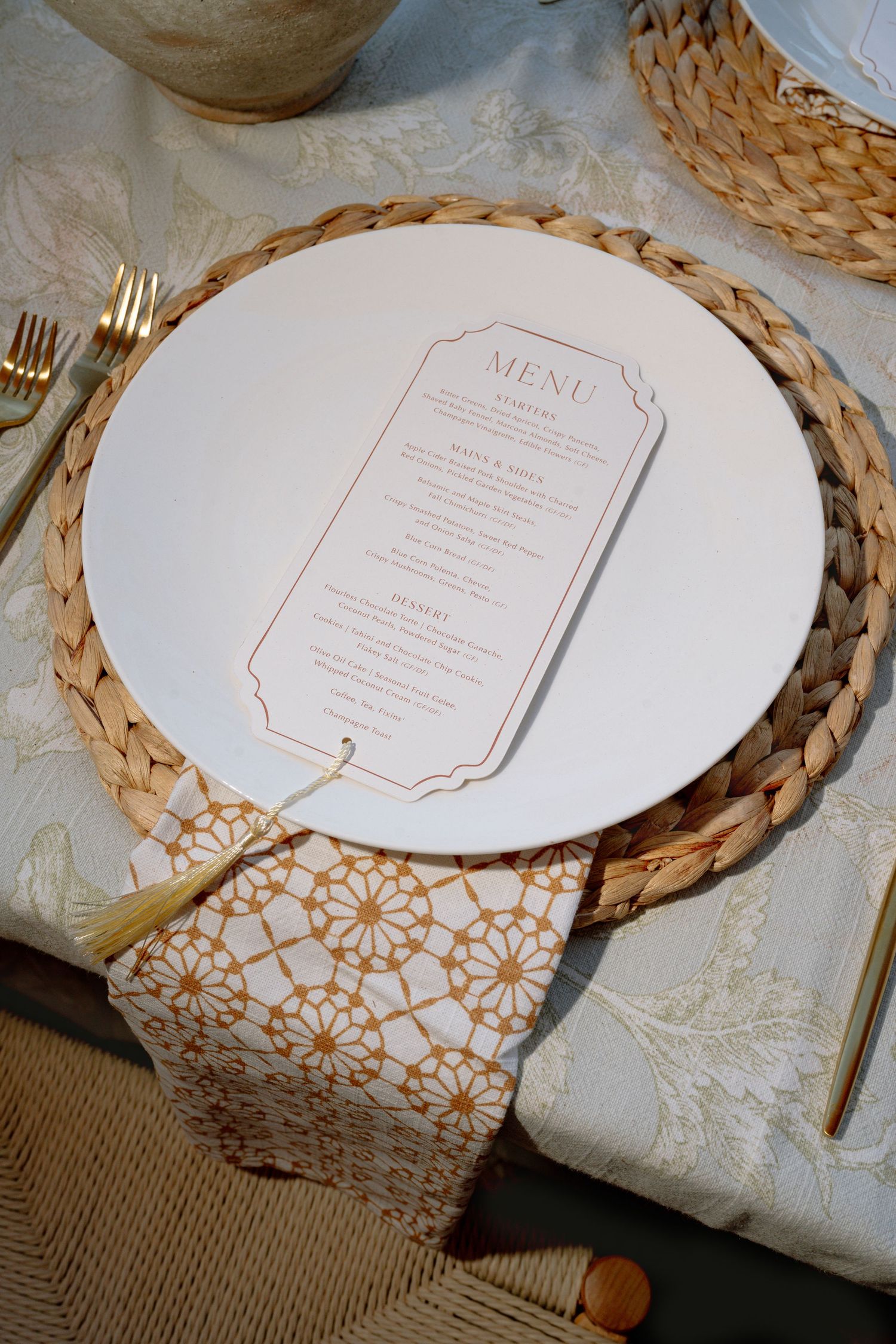 Elegant wedding place setting with white menu card on woven charger, gold flatware, and patterned napkin on linen tablecloth.