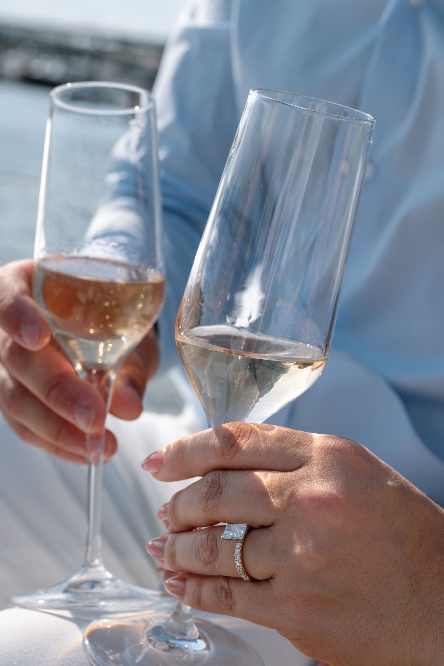 Couple toasting with champagne flutes near water, woman wearing diamond engagement ring, celebrating special occasion.