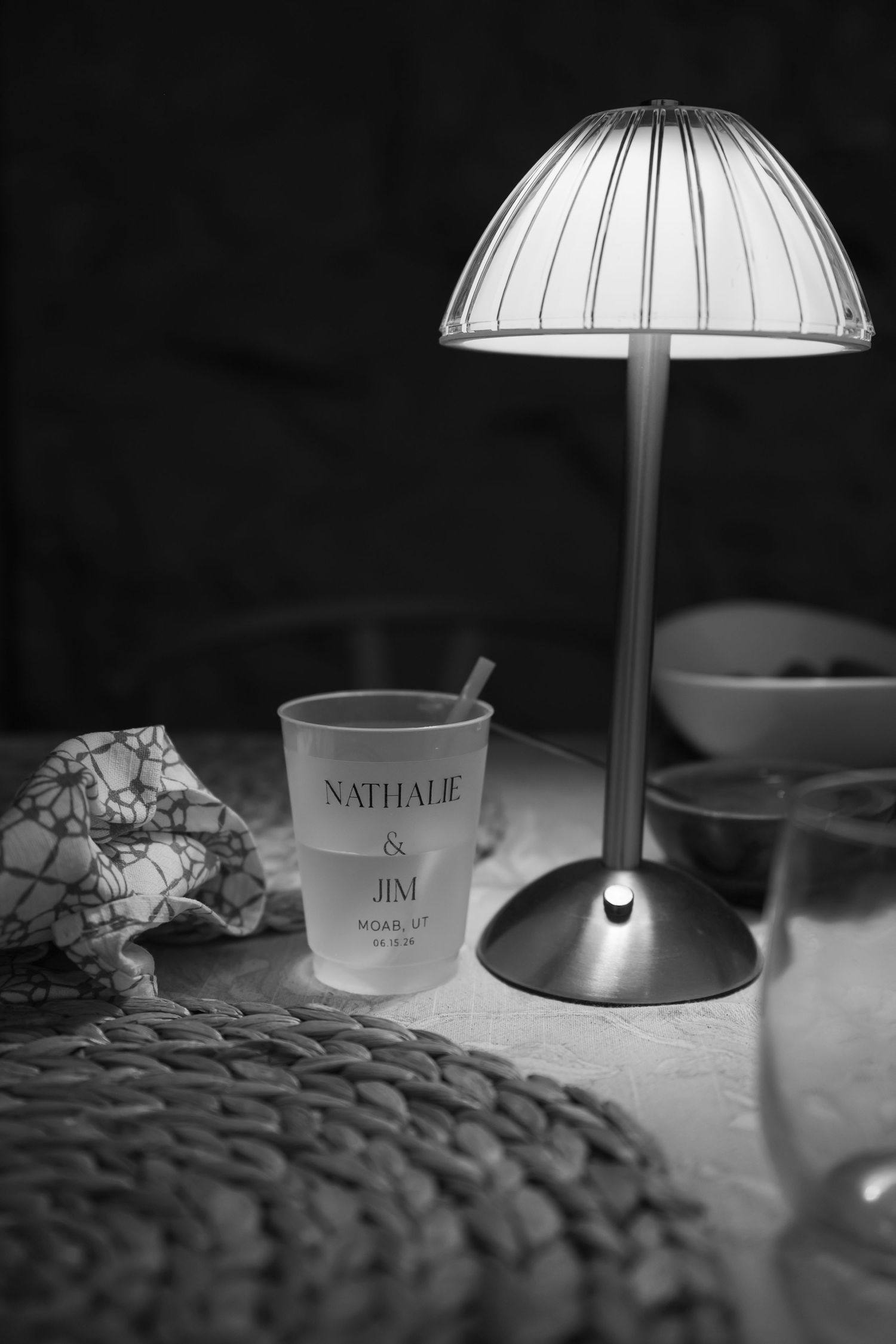 Black and white photo of personalized Nathalie and Jim wedding cup beside elegant table lamp at reception.