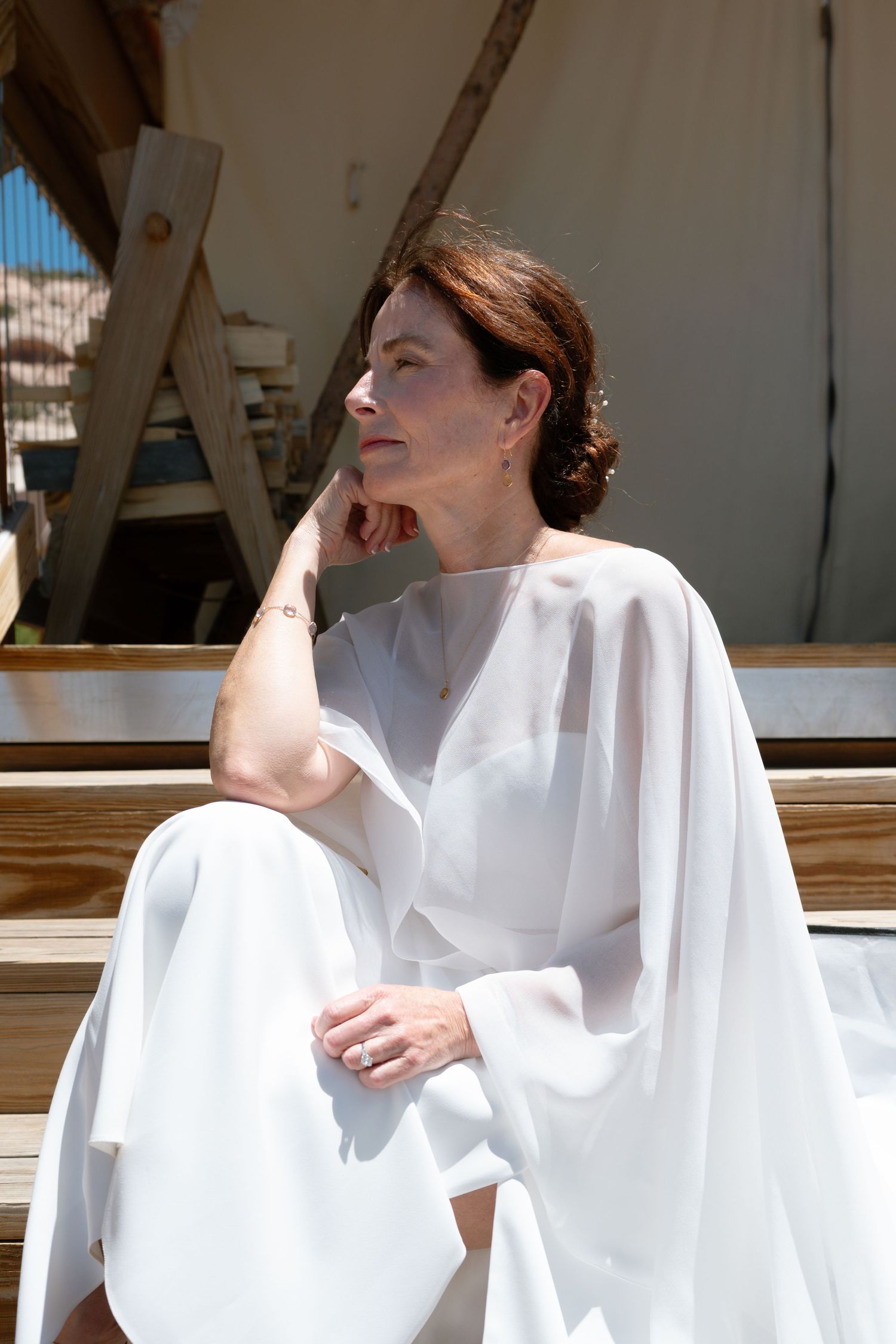 Woman in white flowing outfit sitting on wooden steps, gazing thoughtfully in bright sunlight.