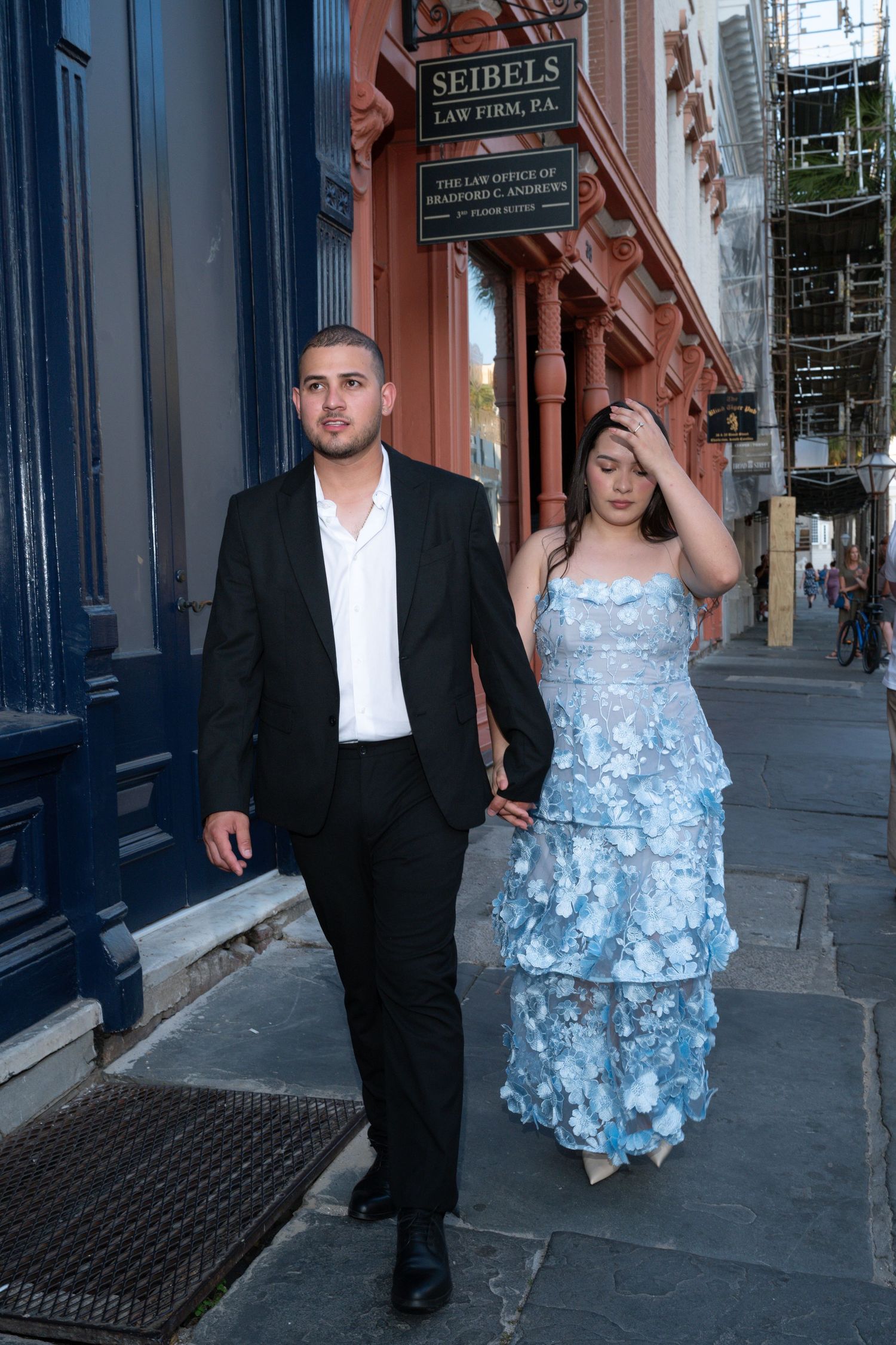 Couple in formal attire walking on a city sidewalk, man in black suit, woman in light blue floral tiered gown.