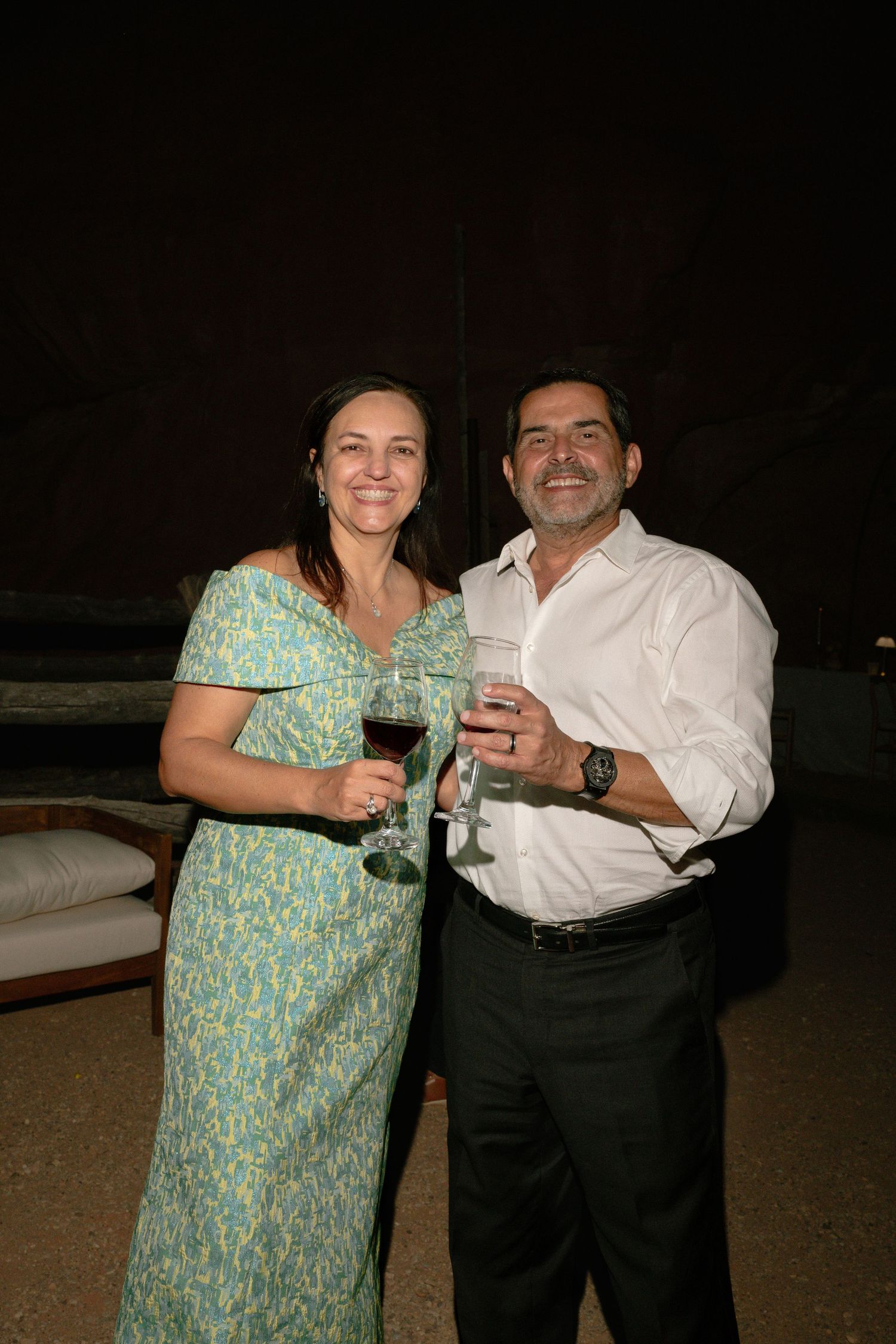 A smiling couple enjoys wine together at an elegant evening event, the woman in a floral dress and man in a white shirt.