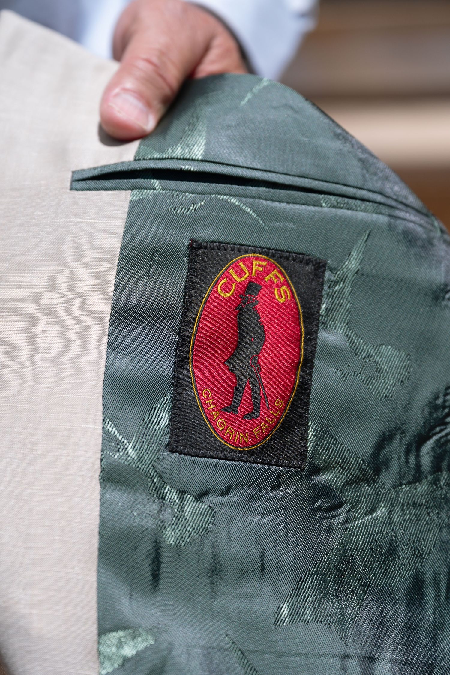 Close-up of a Cuffs Chagrin Falls brand label on the green floral lining of a beige blazer jacket.