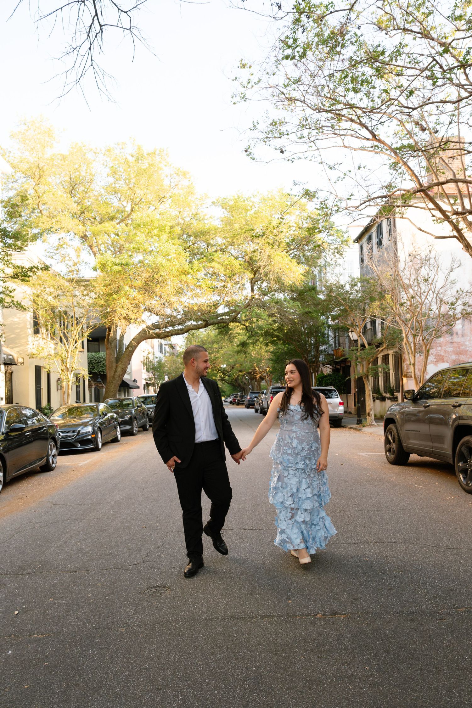Couple holding hands walking down a charming tree-lined street in Charleston, SC, woman in blue floral dress.