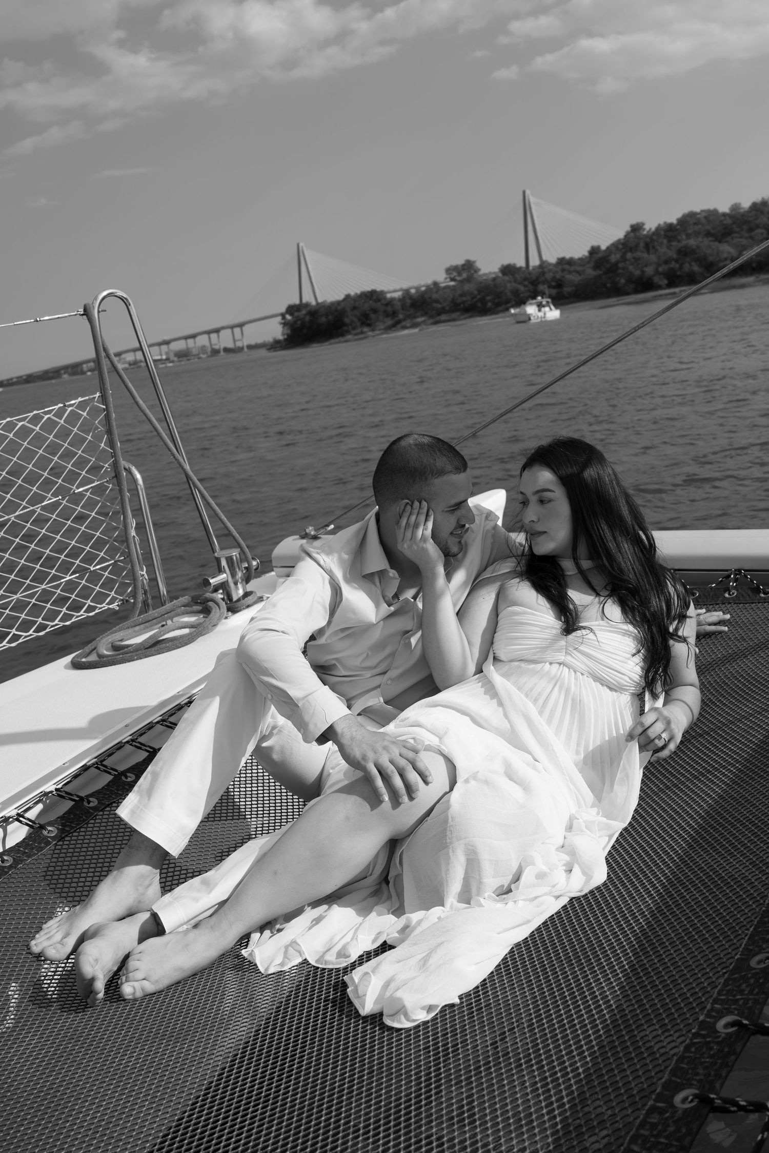 Black and white photo of a romantic couple in white attire relaxing on a sailboat deck with a bridge and river in the background.