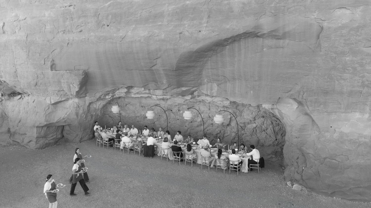 Black and white photo of guests dining at a long table set inside a dramatic sandstone canyon alcove.