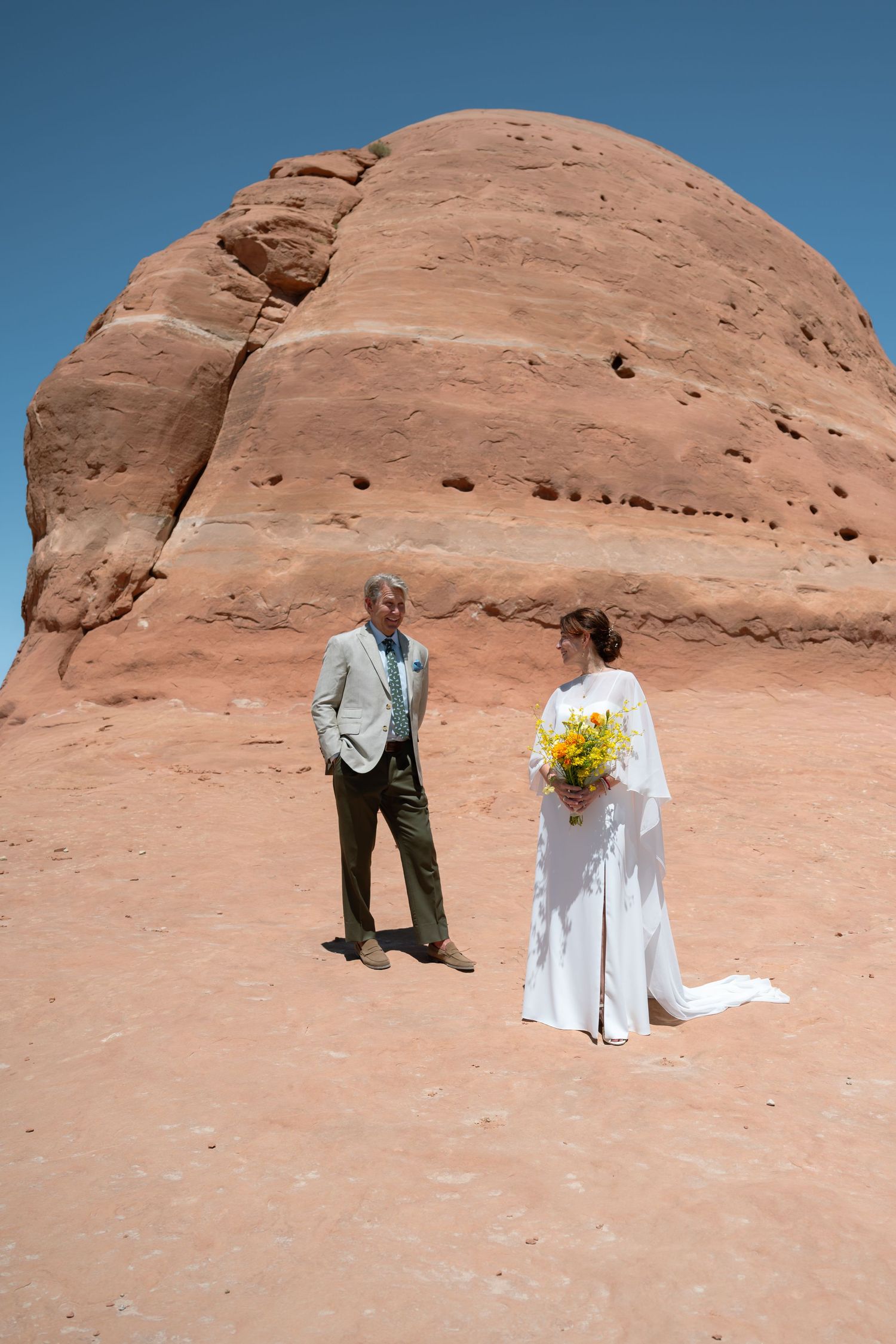 Bride in white gown holding yellow bouquet stands with groom in suit before red sandstone rock formation in desert.